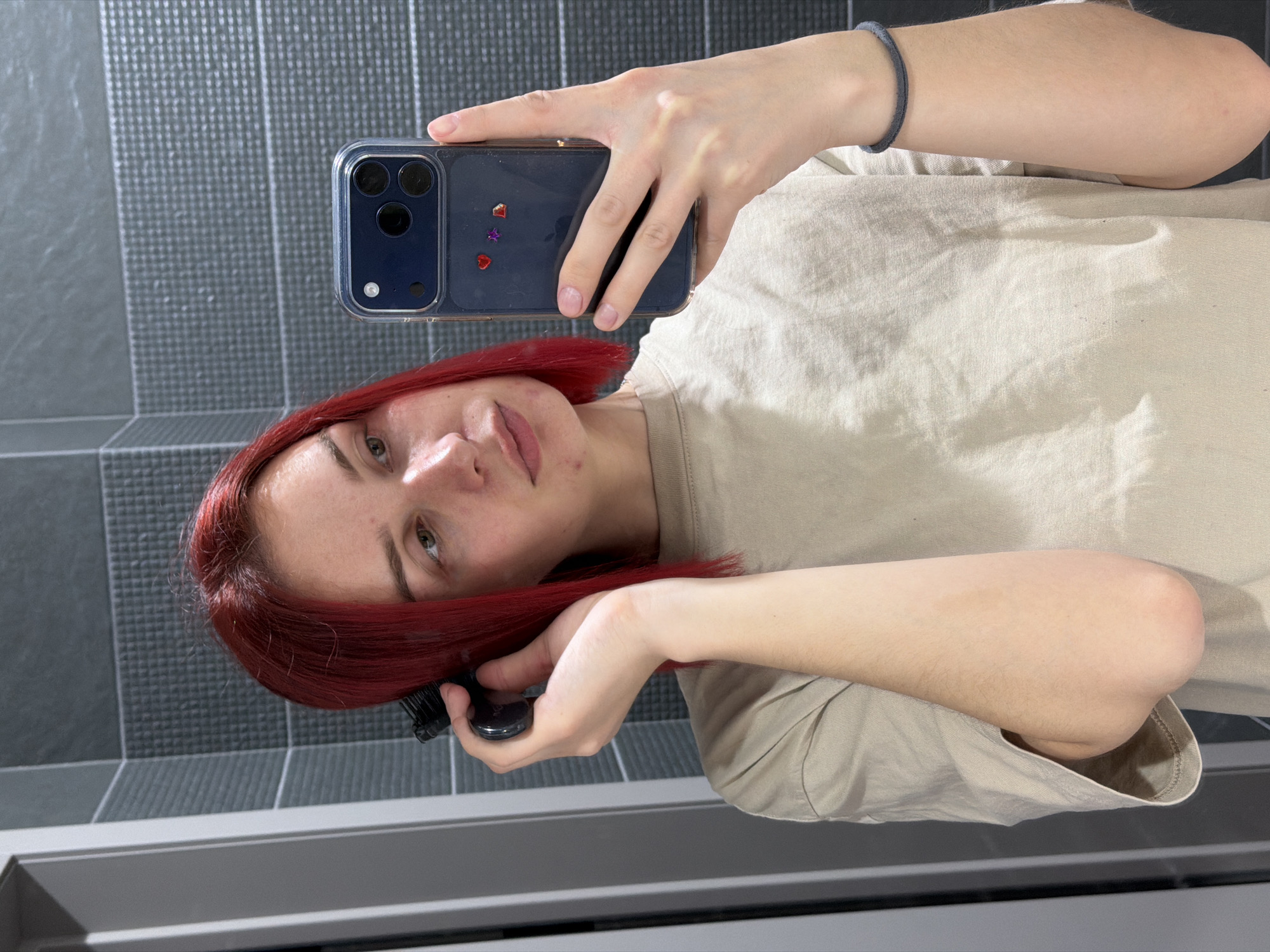 A young woman with red hair is taking a selfie in a bathroom. She is holding a black smartphone in her right hand and using her left hand to hold a black hairbrush near her hair. She is wearing a light brown t-shirt and a dark blue bracelet on her right wrist. The background features gray tiled walls and a horizontal gray element, possibly a countertop or shelf, near the bottom of the frame. The image was taken indoors, likely in a bathroom. There is no information about the specific location in the city of Chaikovsky, Russia.