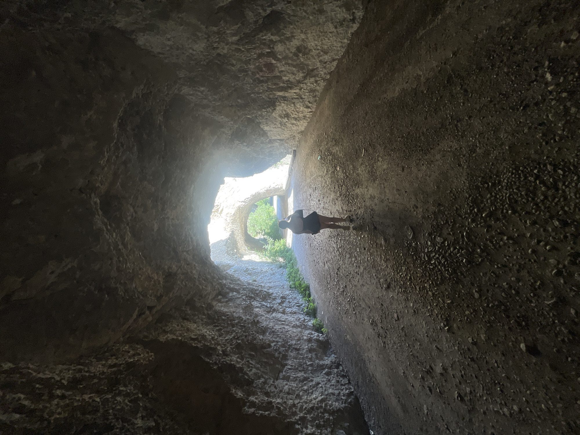 A solitary person, viewed from behind and at a distance, walks away from the camera down a rocky, dirt path within a cavernous tunnel. The tunnel walls are rough and uneven, with the light filtering in from the tunnel's exit ahead. The ground is covered in small stones and dirt. The person appears to be wearing a dark skirt and a light-colored shirt. The scene is located in Asparuhovo, Bulgaria, likely a natural or man-made tunnel in the area.