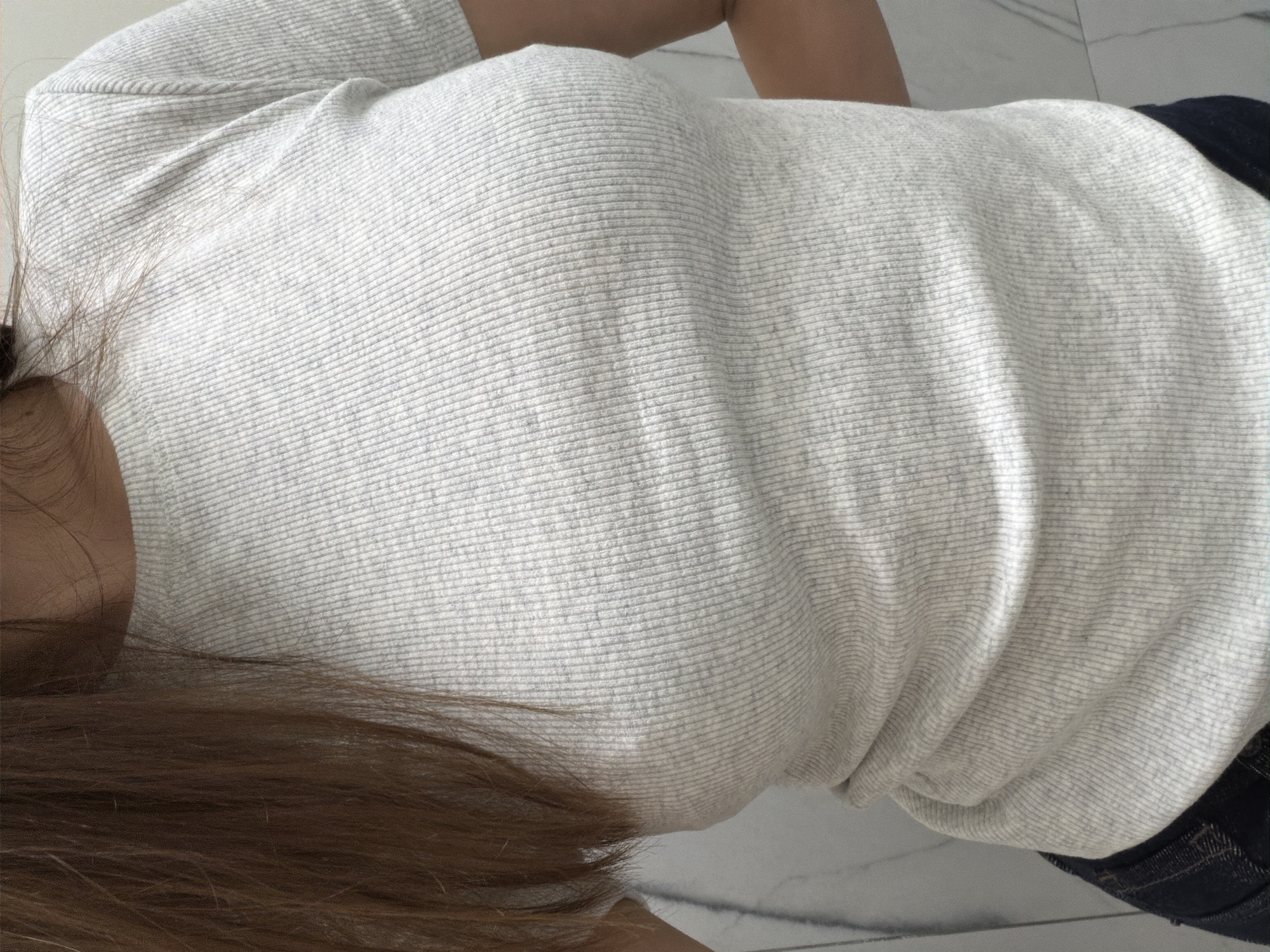 The image shows a medium close-up of a person wearing a light gray, ribbed t-shirt and dark denim jeans. The person's brown hair is visible on the left side of the frame. The background appears to be a wall with a marble-like pattern. No specific location in Khimki, Russia can be determined from the image. The scene is static and does not depict any actions or interactions.