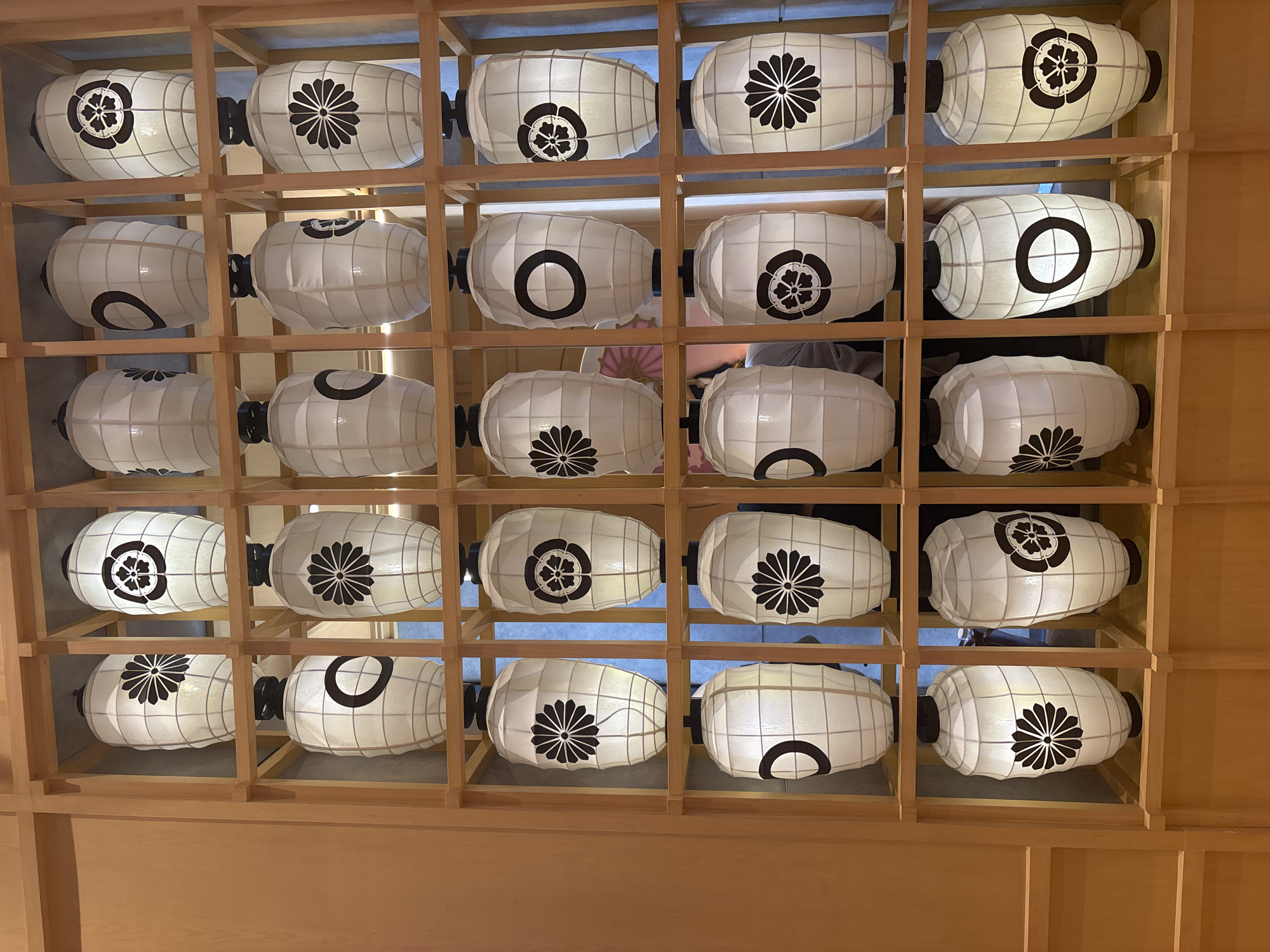 The image displays a wall of vertically oriented, white, paper lanterns, each illuminated from within and featuring a black, traditional Japanese design. These lanterns are organized in a grid of four columns and four rows, framed by light-colored wooden shelving. The designs on the lanterns include a stylized flower with eight petals, a circular motif with a smaller circle inside, and a floral crest within a circle. Some lanterns show variations in the placement and detail of these symbols. The lighting is soft, emanating from the lanterns themselves, creating a warm and inviting atmosphere. The background is out of focus, but appears to be an interior setting with wooden paneling. The scene is located in Kuta, Indonesia, likely within a restaurant or cultural establishment. No people are visible in the image.