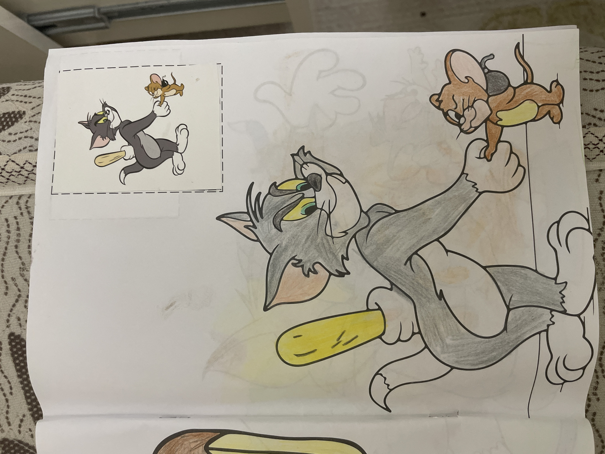 This media file captures a close-up of a coloring page from a coloring book, located in Ihtiman, Bulgaria. The main subject is a partially colored drawing of the iconic cartoon characters, Tom and Jerry.
In the central part of the image, Tom, the gray cat, is depicted with black outlines and colored gray fur, green eyes, and light yellow accents. He is shown actively pursuing Jerry, holding a yellow bat in his right hand. Jerry, the brown mouse, is colored in shades of brown and light yellow, with black features. He is cleverly hanging from Tom's outstretched left hand, appearing to evade Tom's grasp. The larger drawing of Tom and Jerry is still incomplete, with parts of their bodies colored and other areas remaining white outlines.
In the upper left corner of the page, there is a smaller, fully colored reference image of the exact same scene, enclosed by a dashed line, suggesting it's a sticker or a cut-out guide for coloring. The background of the main coloring page features faint, uncolored outlines of other cartoon elements, possibly more hands or abstract shapes, indicating a larger, more complex scene.
The page itself rests on a patterned textile surface, possibly a blanket or a rug, suggesting an indoor and casual setting for this coloring activity. There is no visible text on the coloring page other than the dashed outline around the reference image. The scene captures a dynamic moment of pursuit and evasion characteristic of the Tom and Jerry cartoon.