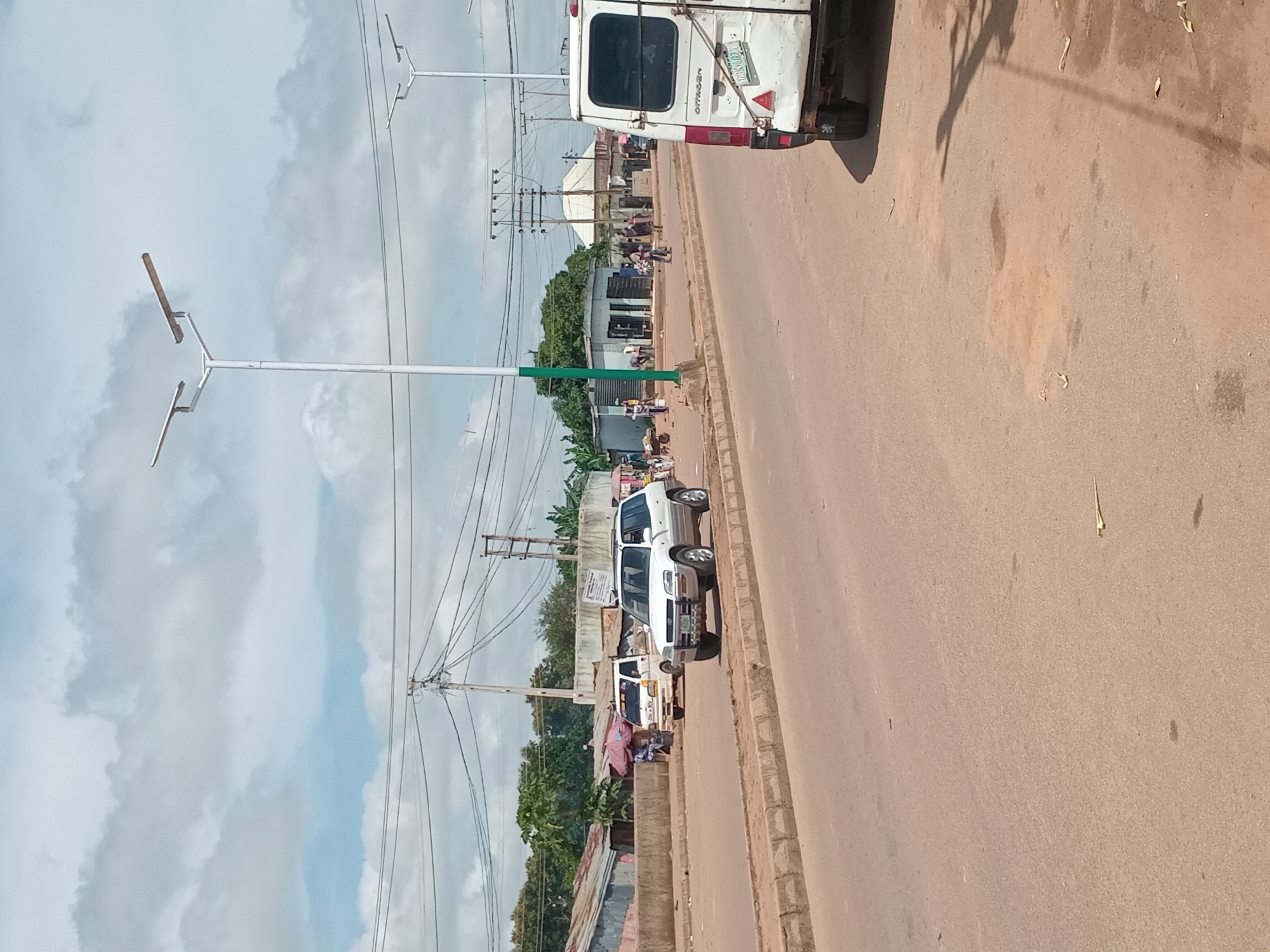 The image shows a street scene in Bukuru, Nigeria. A white SUV is parked on the right side of the road, and a white van is parked on the left. In the background, there are several buildings, including some shops and houses. There are also a few people walking on the street. The sky is cloudy. The road appears to be paved with asphalt, and there are some dirt patches and debris on the side of the road. Power lines and street lights are visible overhead.