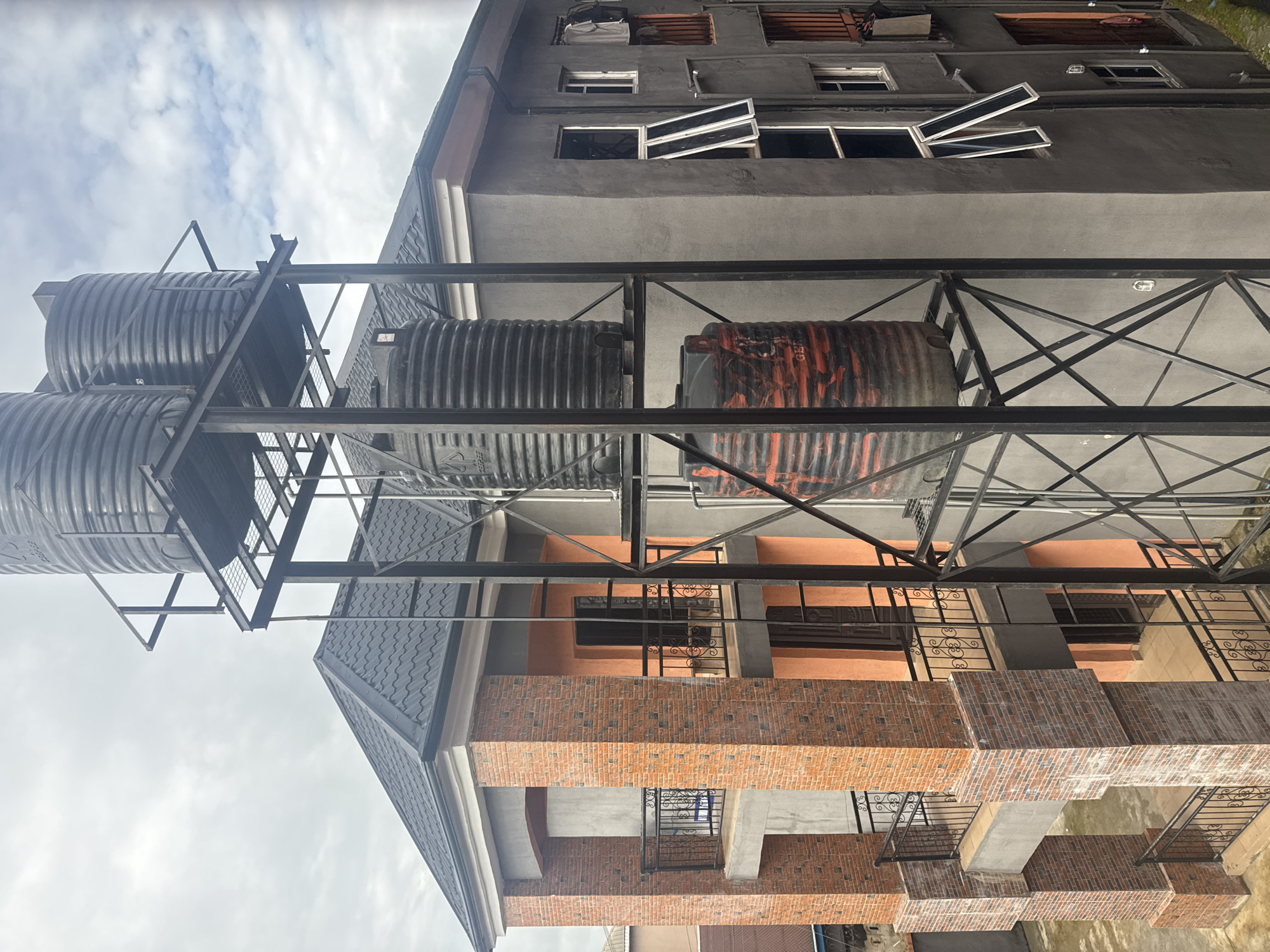 The image captures a close-up, elevated view of a building in Ihiagwa, Nigeria, dominated by a metal water tank structure. Three large black water tanks are mounted on a multi-level black metal scaffolding system. The building itself features a dark grey concrete exterior with several windows, some of which are open. A section of the building is adorned with orange-colored plaster and a brick facade. The roof is made of dark, corrugated material. The sky is overcast with clouds. The scene is static, with no visible human activity or interaction.
