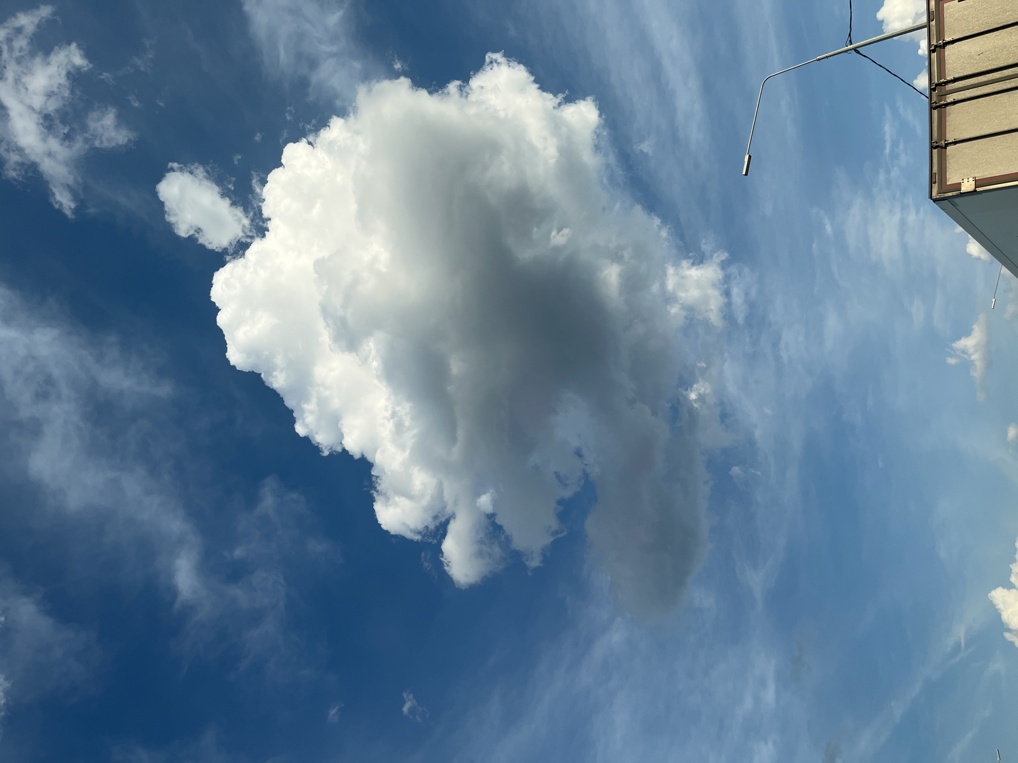 The image depicts a bright, daytime sky with a vibrant blue hue. Large, fluffy white clouds dominate the upper portion of the frame, with a particularly prominent cumulonimbus cloud taking center stage. Thin, wispy cirrus clouds are scattered across the sky, suggesting dynamic weather patterns.
In the lower right corner, a streetlamp with a modern, minimalist design extends upwards, its metallic pole a stark contrast to the organic shapes of the clouds. Below the streetlamp, the top edge of a large, beige cargo truck is visible. The truck's metallic doors and structural details suggest it is a vehicle used for transport. The scene is devoid of any people, focusing entirely on the interplay between the sky, clouds, and man-made elements. The overall impression is one of open space and a clear, though cloudy, day.
