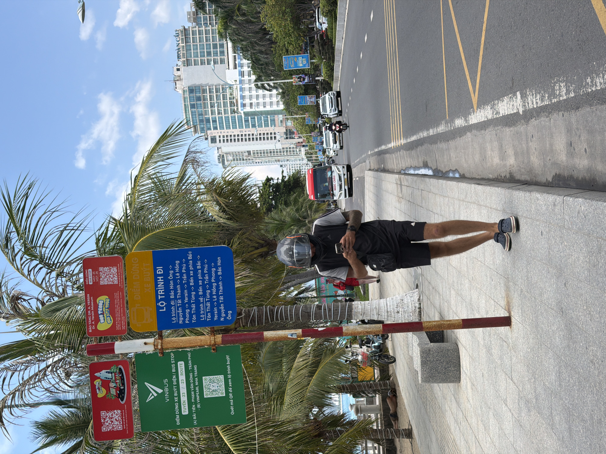 An outdoor urban scene in Nha Trang Ward, Vietnam, depicts a bustling street bordered by a sidewalk lined with palm trees under a clear blue sky with scattered white clouds. The image captures a sunny daytime moment.
In the foreground, a man wearing a black t-shirt, black shorts with white stripes, a dark helmet with a clear visor, and dark sandals stands on the grey tiled sidewalk. He is engrossed in his mobile phone, held in both hands, and also has a black fanny pack or small bag across his chest. Further down the sidewalk, partially obscured by palm trees, another person with a bare torso is visible.
Next to the man stands a striped pole (red, white, and brown) featuring several informational signs. A red sign indicates a "BUS STOP" for the "Dr. A. Yersin Museum" with route numbers (MST: 25, MST: 26) and QR codes. A blue sign, titled "ĐIỂM DỪNG XE BUÝT" (BUS STOP), details "LỘ TRÌNH ĐI" (Route to) and "LỘ TRÌNH VỀ" (Return route) with various locations including "Bắc Hòn Ông", "Nguyễn Tất Thành", "Lê Hồng Phong", "Yersin", "Trần Phú", "Tôn Thất Tùng", and "Bến xe phía Bắc". Below it, a green "VINBUS" sign advertises an "ĐIỂM DỪNG XE BUÝT ĐIỆN / BUS STOP" for "TUYẾN 23 / 23 ROUTE" connecting "(A) BÃI TIÊN - PHẠM VĂN ĐỒNG - TRẦN PHÚ" to "(B) VINPEARL NHA TRANG", urging users to "Quét mã QR để xem lộ trình buýt!".
Behind the sidewalk, a street with yellow markings features a concrete curb. Vehicles, including a red and white bus marked "NGỌC CHÂU", white cars, and motorcycles, are visible moving along the road. In the background, tall, modern buildings rise, one clearly displaying "HOÀN CẦU". Streetlights are also visible along the road.