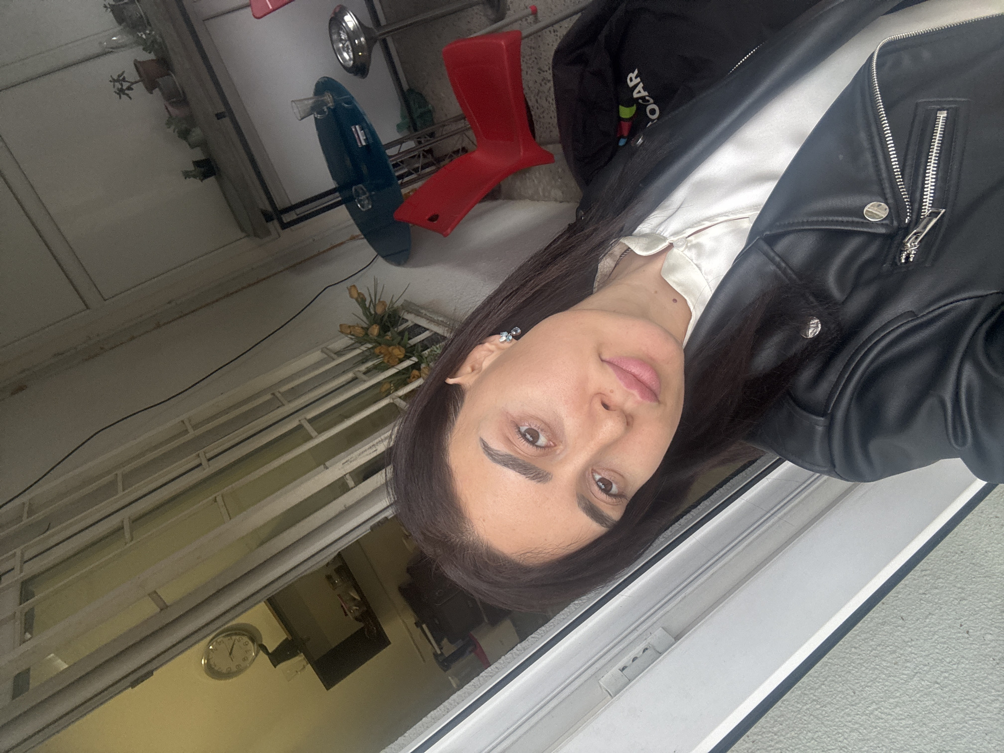 A woman wearing a black leather jacket and a white satin shirt is positioned near a window in a building in Tbilisi, Georgia. Behind her are interior elements like a red stool, a blue table, and a shelf with potted plants. A clock is visible on the wall.