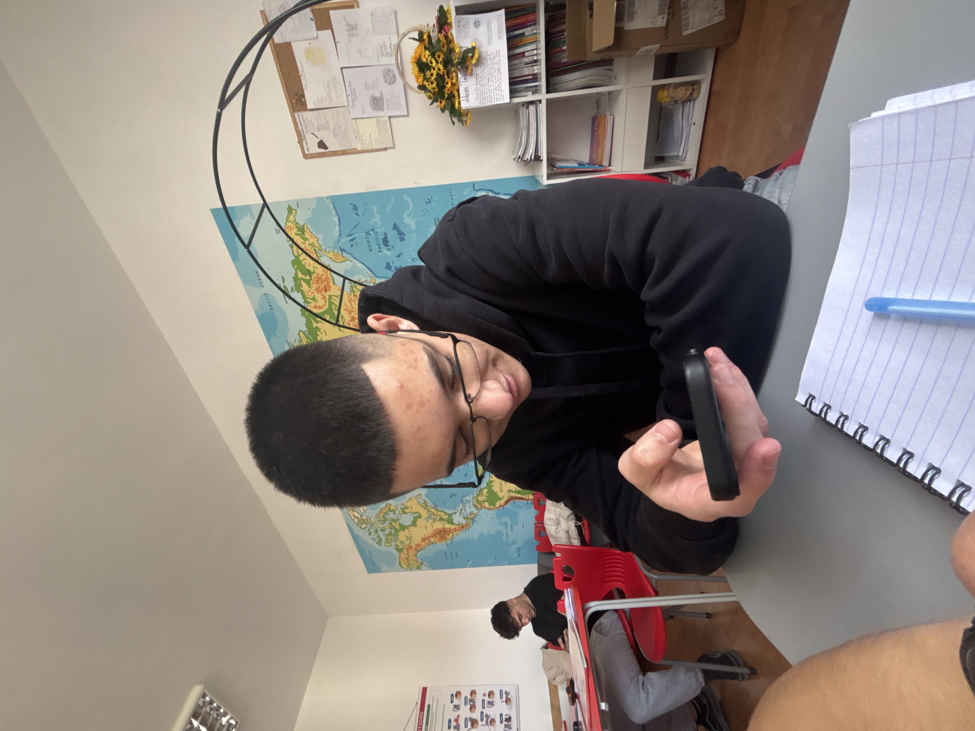 A young man with short dark hair and clear-framed glasses, wearing a black hoodie, is the central figure in an indoor setting that appears to be a classroom or study area. He is seated at a gray desk, looking intently at a black smartphone held in his hands. His expression is focused on the device.
In the background, a large world map adorns the wall, displaying geographical features including parts of North America and Asia, with "NORTH PACIFIC OCEAN" and "ARCTIC OCEAN" clearly visible. Above the map, a cork board holds various papers, one of which has "Mem." discernible. A decorative black metal hoop structure is partially visible near the cork board. Adjacent to these, white shelves are stocked with books, folders, and boxes. A small cluster of artificial yellow flowers, resembling sunflowers, adds a decorative touch.
To the right of the main subject, a white spiral-bound notebook with blue ruled lines is open on the desk, with a blue pen resting on it. In the mid-ground, another young man with dark hair is seated at a separate desk with a red-backed chair, his head bowed, seemingly engaged in an activity or study. Part of a person's arm or leg is visible in the lower right foreground. Near the bottom left, a partially visible red sign or poster shows the text "TIONS".
The lighting suggests it is daytime. The overall scene depicts individuals in a learning or working environment. The location is specified as Zlatograd, Bulgaria.