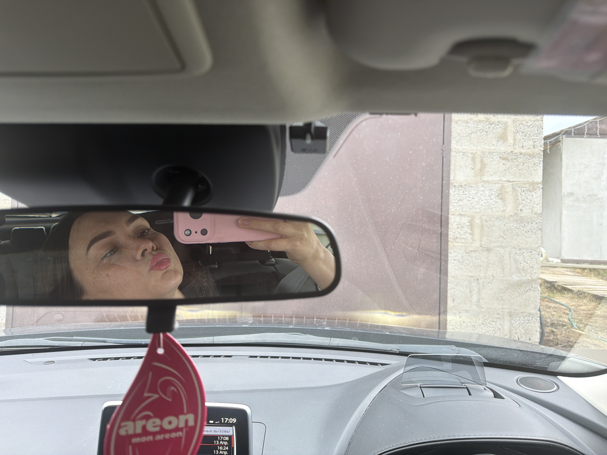 A photograph taken from the interior of a car shows a woman reflected in the rearview mirror. The woman is holding a pink phone up to the mirror, presumably taking a selfie. She is making a kissing face towards the camera.
In the background, visible through the car's windshield, is a building exterior constructed of cinder blocks. The windshield appears to be slightly dirty. A dark-colored garage door is partially visible on the left side of the frame. A head-up display (HUD) system is located on the car's dashboard. An air freshener is hanging from the rearview mirror, partially obstructing the driver's view. The car's instrument panel shows a timestamp of 17:09 and several dates, indicating the image was taken on April 13, 2024.
The location is Chaikovsky, Russia.