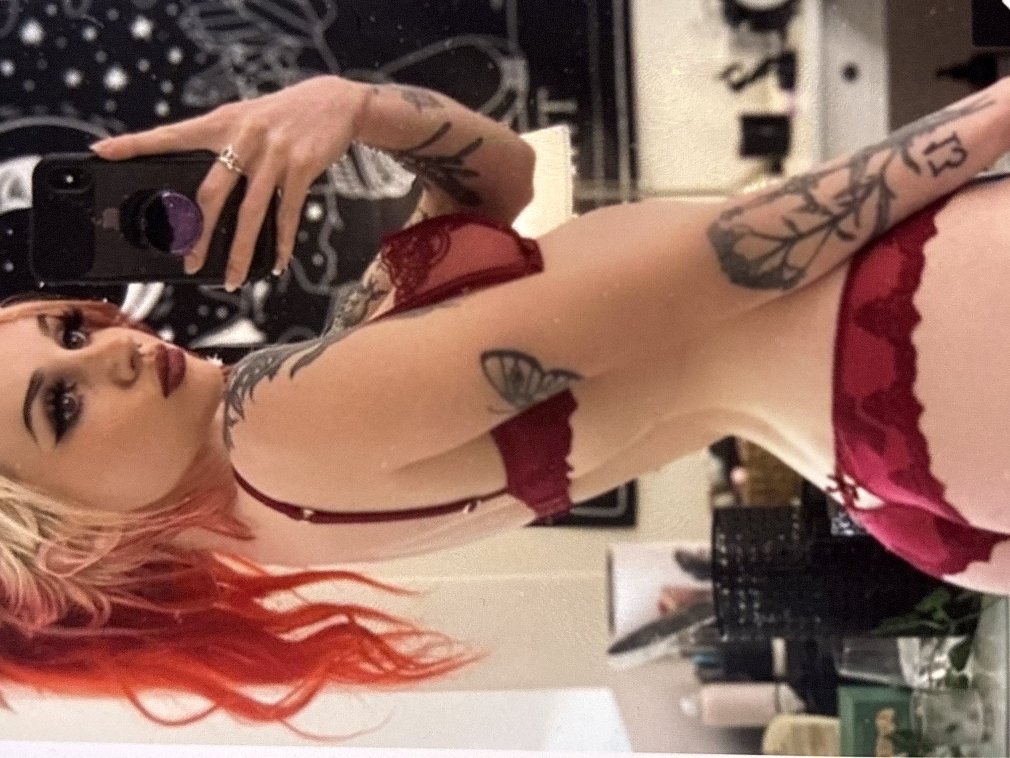 A person with brightly colored red and blonde hair, wearing a red lace bra and matching underwear, poses for a mirror selfie. Their right hand holds a smartphone with a purple pop socket, and a ring is visible on their ring finger. Tattoos are visible on their arms and shoulder. The background shows a cluttered dresser with various items, including bottles, jars, and a black basket. A tapestry with a celestial pattern hangs on the wall behind them. The lighting suggests an indoor setting, likely a bedroom or bathroom.