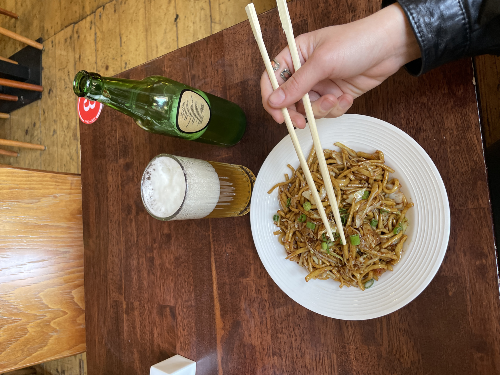 An overhead shot captures a casual dining scene on a dark wooden table in Tbilisi, Georgia. A person's fair-skinned right hand, adorned with two rings, grips a pair of light wooden chopsticks, poised to pick up noodles from a white, fluted plate. The person wears a black leather jacket, with a cuff visible at the wrist.
The plate is filled with stir-fried noodles, likely lo mein or chow mein, mixed with shredded cabbage or sprouts and garnished with chopped green onions. Beside the plate, a clear glass mug holds a light-colored beer with a significant frothy head. Further to the left, a dark green glass bottle lies horizontally, with a brown label on its side. The label clearly shows Georgian script, reading "მუშმალა" (Mushmala), which refers to a medlar or loquat fruit, and a date "26.07.2020". A red circular sticker with a white numeral "3" is partially visible beneath the bottle's neck.
The table itself is polished wood, reflecting some light. To the far left, a section of a lighter wooden surface, possibly a different table or a wall panel with horizontal wooden slats, is visible. The scene suggests an indoor setting, likely a restaurant or bar, focused on a meal of Asian-inspired noodles and a beverage.