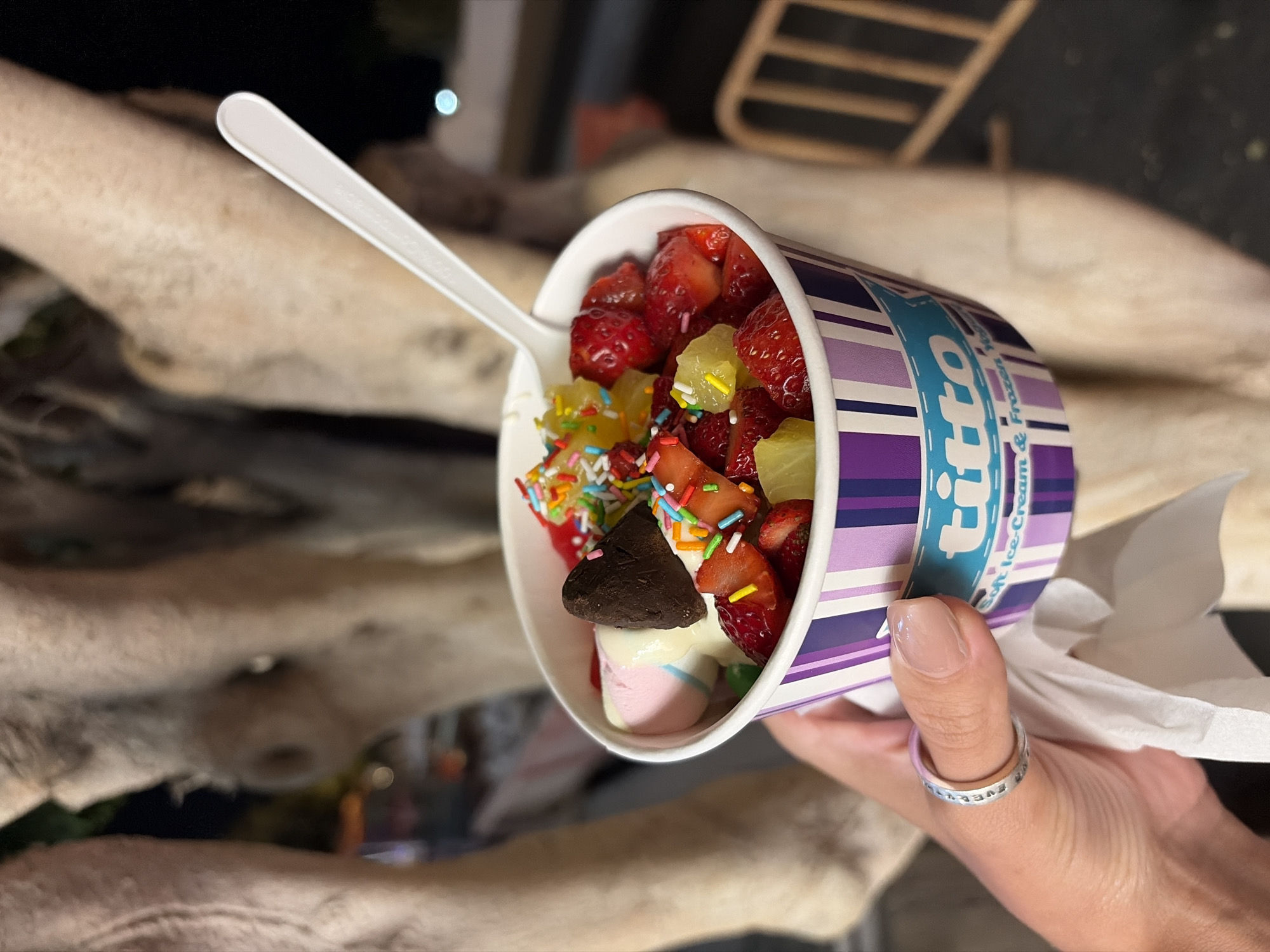 A close-up, slightly tilted image shows a person's hand holding a colorful cup of frozen yogurt or soft-serve ice cream, viewed from a slightly elevated angle. The cup features vertical stripes in shades of purple and white, with a teal banner displaying the brand name "fitto" and the text "Soft Ice Cream & Frozen Yogurt" below it.
The dessert is generously topped with an array of fresh fruits and confectionery. Visible toppings include bright red chopped strawberries, yellow pineapple chunks, vibrant multi-colored sprinkles, a dark chocolate-covered piece (possibly a cookie or brownie) shaped like a heart, and soft pink and white marshmallows. A white plastic spoon is inserted into the cup, with faint, unreadable text on its handle.
The hand holding the cup has a light, natural-colored nail polish and wears a silver-toned ring on the thumb, partially engraved with text that appears to say "LIVE EVERY". A crumpled white napkin or tissue is visible beneath the cup.
The background is highly out-of-focus, suggesting a shallow depth of field. It features large, light-colored, rounded shapes that could be natural rock formations or sculpted concrete, characteristic of an outdoor setting. Blurred lights in the distance indicate that the scene takes place in the evening or at night. The overall impression is one of a leisurely moment enjoying a sweet treat in an outdoor setting, potentially a coastal area or promenade in Puerto Del Carmen, Spain.