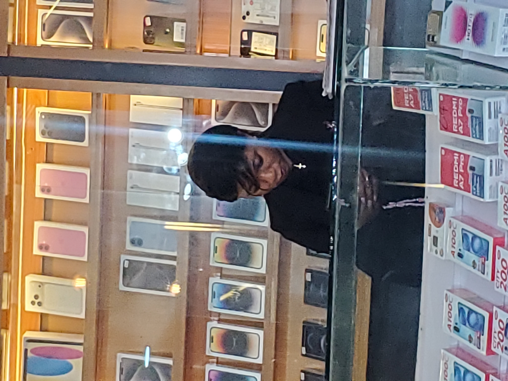 This media file depicts a person, likely a shopkeeper, standing behind a display of mobile phones. The shop appears to be a retail establishment, possibly a phone store, with shelves filled with various mobile phone boxes and some individual phones on display. The person is wearing dark clothing and has their head bowed, looking down at the phones. The lighting in the shop is bright, suggesting it is daytime or the shop is well-lit. Reflections on the glass display case indicate that the photo was taken from the outside, looking in. Some visible text on the phone packaging includes "REDMI A7 Pro", "HOT 70", and "CITY 200", which are likely model names or product codes. The overall scene suggests a commercial environment focused on the sale of mobile technology. The location context of Jos, Nigeria, implies this is a typical phone shop within that city. The image's orientation is slightly tilted, but the content remains clear.