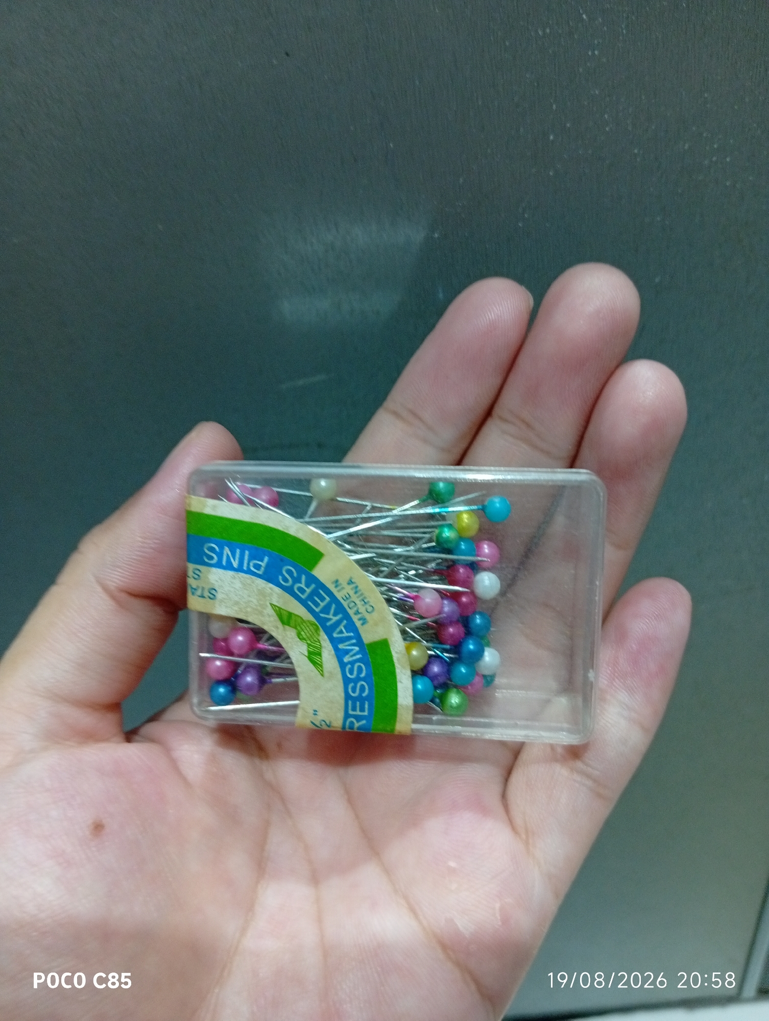 A hand holds a small, clear plastic box filled with straight pins with colorful, spherical heads. The pins are silver with heads in shades of pink, purple, blue, green, and yellow. A label is attached to the box, partially obscuring the pins. The label is green and yellow with text that reads "PRESSMAKERS PINS" and "MADE IN CHINA". The background is a plain, grey textured surface. The timestamp "19/08/2026 20:58" and "POCO C85" are visible in the bottom right and bottom left corners respectively. The scene is likely indoors, given the lighting and background. There is no indication of the specific location within South Tangerang, Indonesia.
