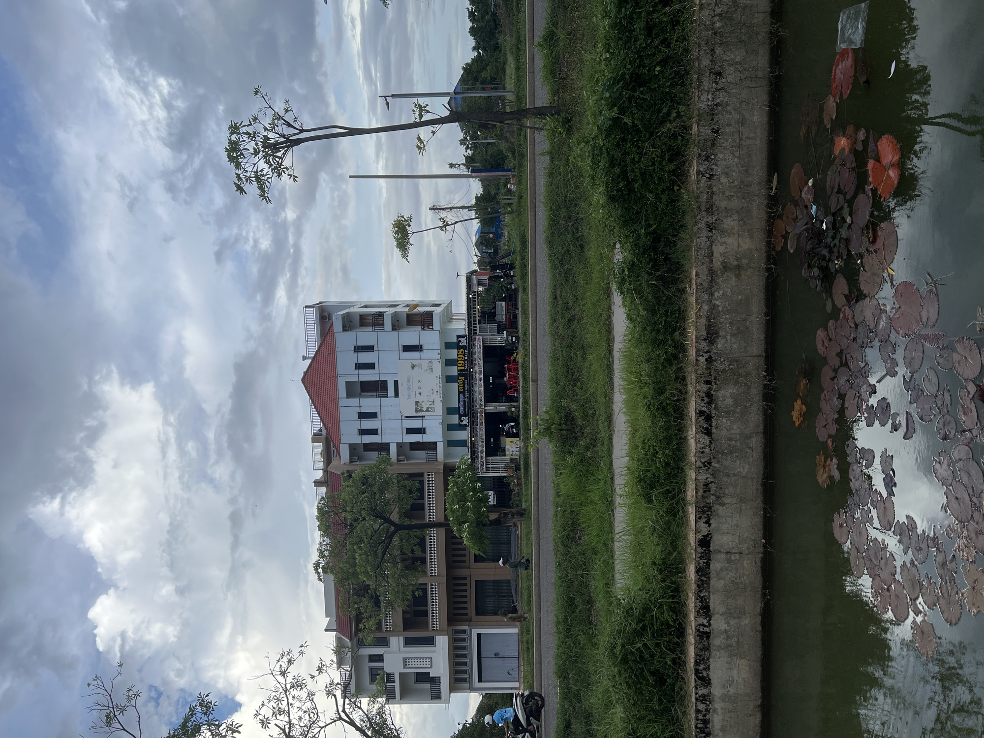 This is an outdoor, daytime, eye-level medium shot of a street scene in Siem Reap, Cambodia. The scene is dominated by a three-story building with a red-tiled roof, featuring a mix of modern architecture with white and beige facades, and balconies on the upper floors. Below the residential floors, commercial establishments with glass storefronts are visible, including one with a sign that reads "1998 Siem Reap". A paved road runs in front of the building, with a motorcyclist wearing a blue shirt and white helmet riding on the left side of the road. Two pedestrians, one wearing a white cap and the other a dark shirt, are walking on the sidewalk. Green grass and small trees line the side of the road. In the foreground, a canal with dark water contains numerous lily pads, some of which are reddish-brown. The sky is cloudy with patches of blue.