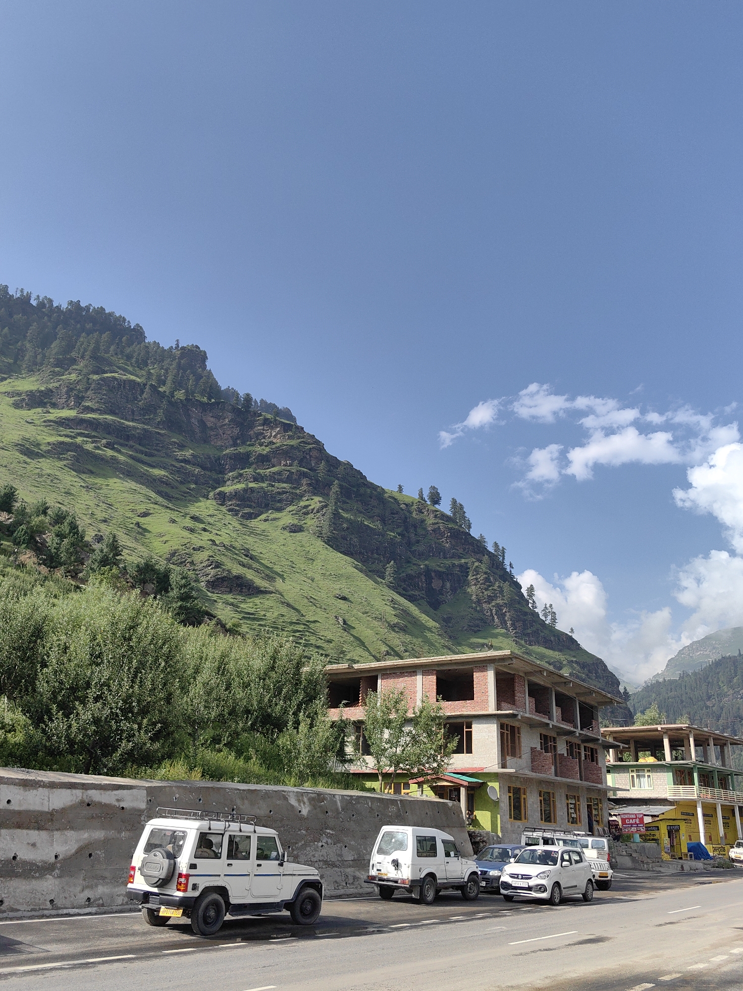 The image depicts a street scene in Palchan, India, with a mountain landscape in the background. In the foreground, several vehicles are parked and driving on a paved road. A white SUV with a roof rack is on the left, followed by a blue car and a white hatchback. Behind them, a white van is visible.
Several buildings line the right side of the road. The closest building is under construction, made of brick and concrete, with unfinished upper floors. Next to it is a finished building with a green facade on the lower level and balconies on the upper floors. Further down the street, a yellow building with a sign that reads "MONTANA TOP CAFE" is visible, suggesting a commercial area.
The environment is mountainous, with a steep, green hillside covered in trees and rocks dominating the left side of the image. The sky is clear blue with a few scattered white clouds. The overall impression is a roadside view in a mountainous region, likely a tourist or transit area given the presence of cafes and ongoing construction. No people are clearly discernible as actively interacting with each other or engaged in specific actions.