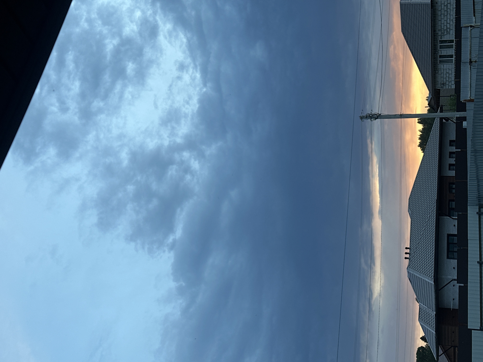 The image shows a landscape with a cloudy sky dominating the upper portion of the frame. The sky transitions from lighter, brighter clouds to darker, more ominous-looking clouds. Power lines run across the middle of the image, and on the right side, there are houses with roofs, some of which appear to be constructed of brick. Some areas between the houses and sky are lit with the colors of sunset. A power pole stands in line with the houses. The image is taken from a location that appears to be a residential area. The city of Kharnavy, Russia is identified as the location. There are no visible people or actions.