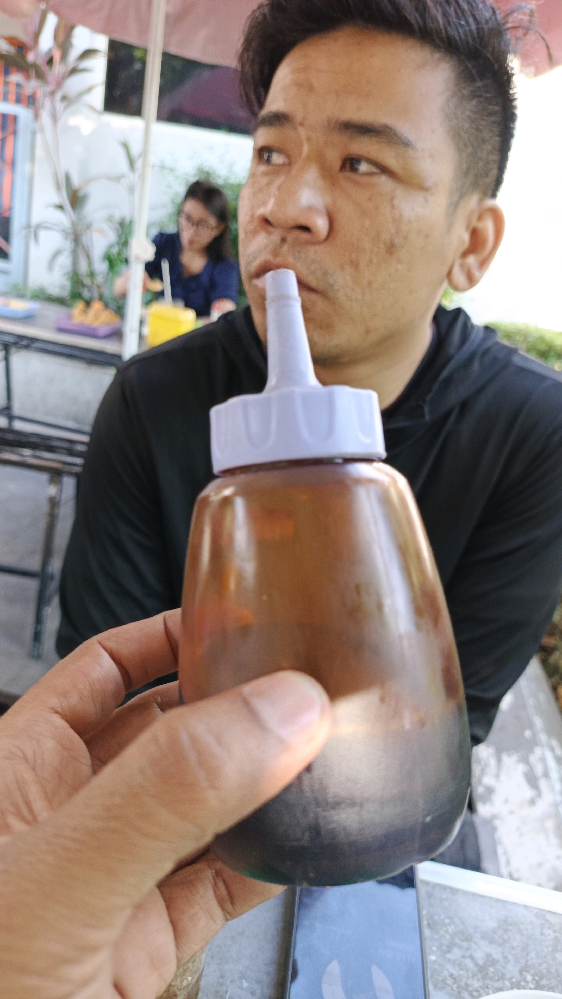 A close-up shot captures a man holding a brown sauce bottle with a white nozzle to his mouth, as if to drink from it, with his right hand in the foreground. The man, facing slightly to the right, has dark hair and is wearing a black hooded shirt. Behind him, a woman with glasses and a dark blue shirt is visible in the background, seated at a table with yellow and white containers. The scene appears to be outdoors, possibly at a restaurant or food stall, with a pink umbrella and foliage suggesting a tropical setting. The presence of a cell phone on the table in front of the man, as well as the overall ambiance, points to a casual dining environment in the city of Surakarta, Indonesia.