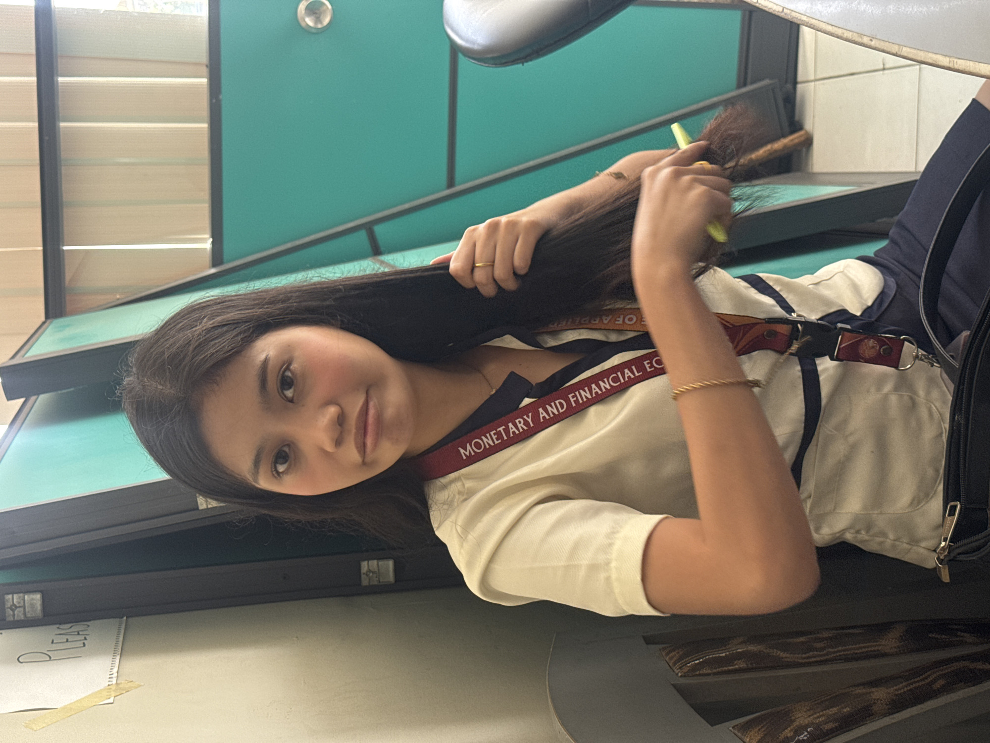 A young Asian woman with dark hair is seated, facing forward, and brushing her hair with a light green brush. She is wearing a white collared shirt with a dark blue trim and a lanyard with the text "MONETARY AND FINANCIAL EC." She has a thin gold bracelet on her left wrist and a ring on her right ring finger. The background is out of focus, but it appears to be an indoor setting with teal partition walls and some vertical blinds. A sign that says "PLEASE" is visible on the left wall, taped with yellow tape. The overall impression is a candid shot of the woman grooming herself in what could be a public or institutional setting.