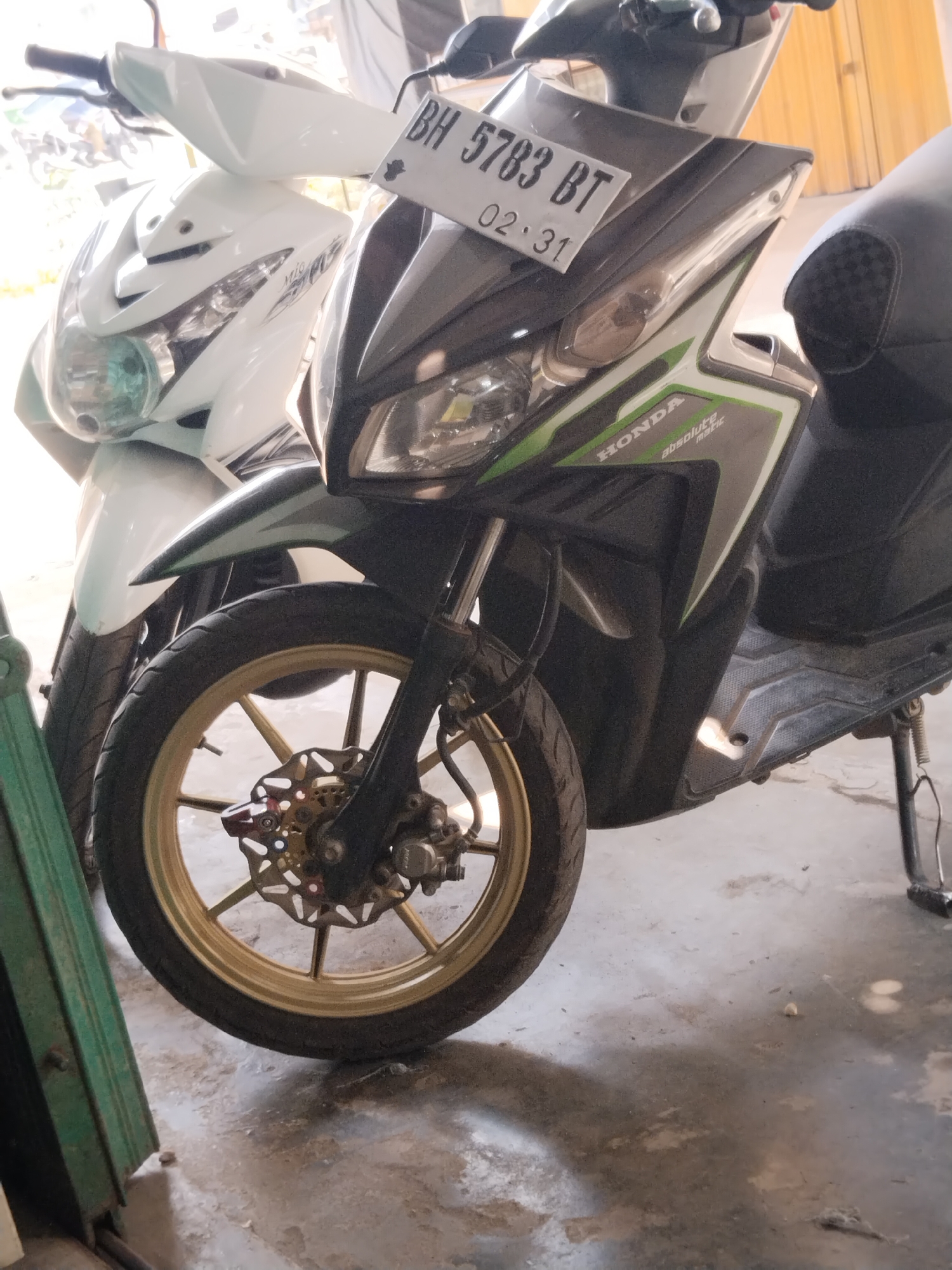 Two parked scooters are shown in a garage or workshop. The scooter in the foreground is black and green and has a gold rimmed wheel with a disc brake. The license plate reads "BH 5783 BT 02.31". The scooter in the background is white. Both scooters appear to be in good condition. The floor is concrete. The lighting is bright, suggesting it is daytime.