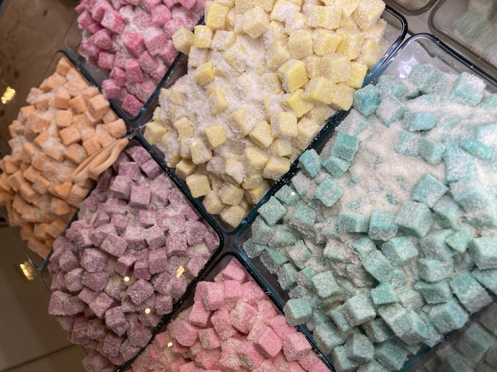 The image shows a close-up, overhead view of several square glass dishes filled with colorful, bite-sized Turkish delight confections. The treats are cube-shaped and coated in shredded coconut. From top left to bottom right, the colors are: light orange, pink, light purple, yellow, and a bright turquoise or teal. The dishes are arranged in a grid-like pattern, creating a visually appealing display of sweets. The background is out of focus but appears to be a reflective surface, possibly a glass counter or display case. There are no people or identifiable landmarks present in the image, and the setting is not specified to be in Barcelona, Spain.