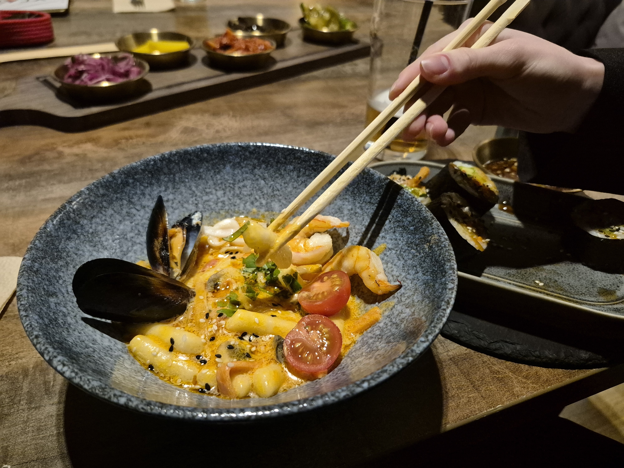 The image shows a person using chopsticks to pick up food from a blue-grey speckled bowl containing pasta with shrimp, mussels, and halved cherry tomatoes in a light orange sauce with herbs and sesame seeds. To the right of the bowl, there's a dark rectangular plate with what appears to be sushi. A dark-colored sauce is also visible on that plate.
Behind the bowl and sushi, there is a wooden tray holding several small dishes with different types of condiments. In the background, there appears to be a glass with liquid inside. The scene is set on a wooden table.
There is no verifiable evidence the image was taken in Chaikovsky, Russia, or any other location.