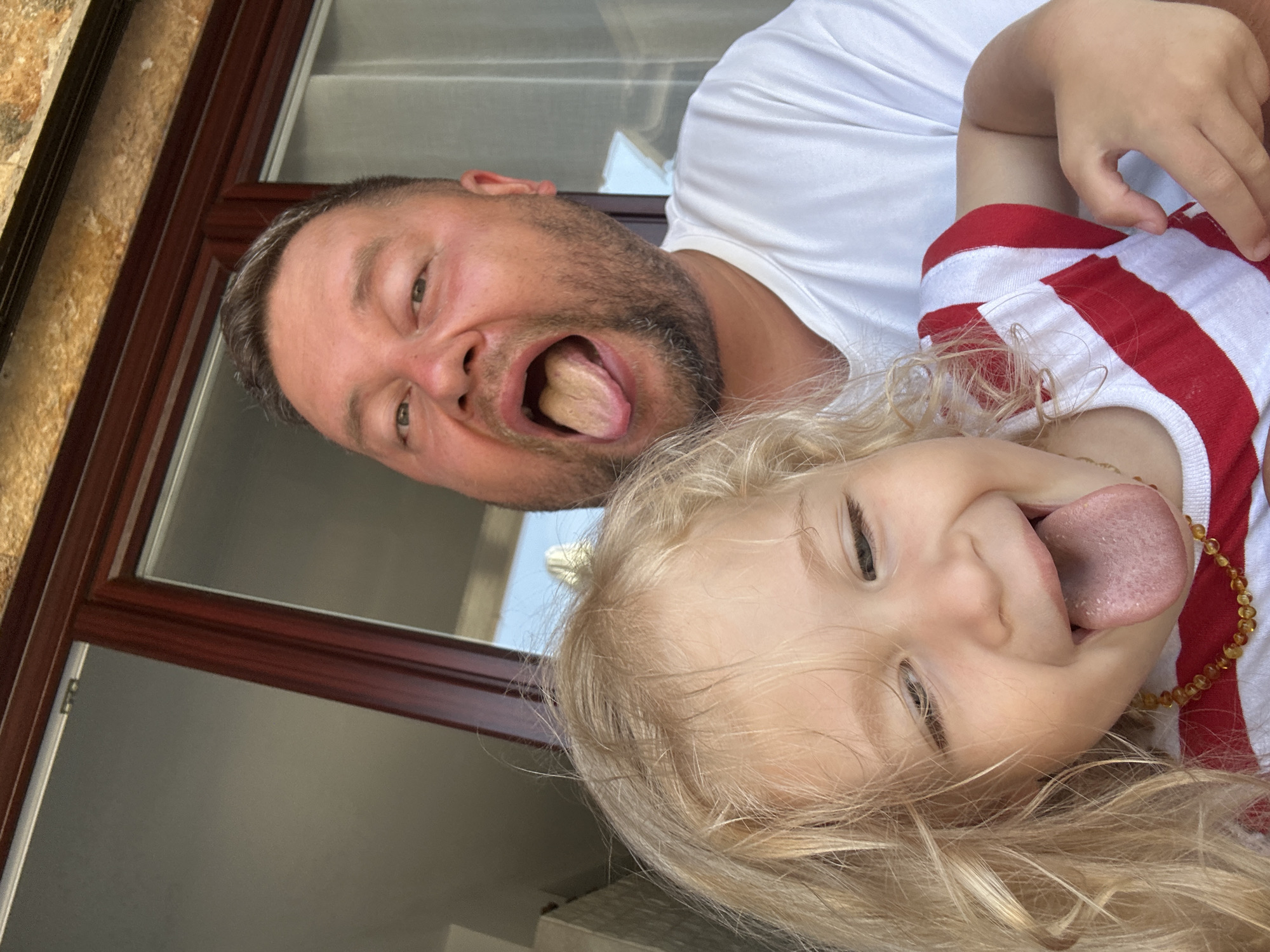 The image shows a close-up selfie of a man and a young child, likely father and daughter, with their tongues sticking out. They are both wearing white t-shirts, the child's also has red stripes. The man has short, dark hair and a beard. The child has blonde, curly hair. They appear to be outdoors, possibly on a balcony or near a window, with a blurry background that includes a building and some greenery. The lighting suggests it is daytime. The image was taken in the city of Sant Feliu de Guíxols, Spain.