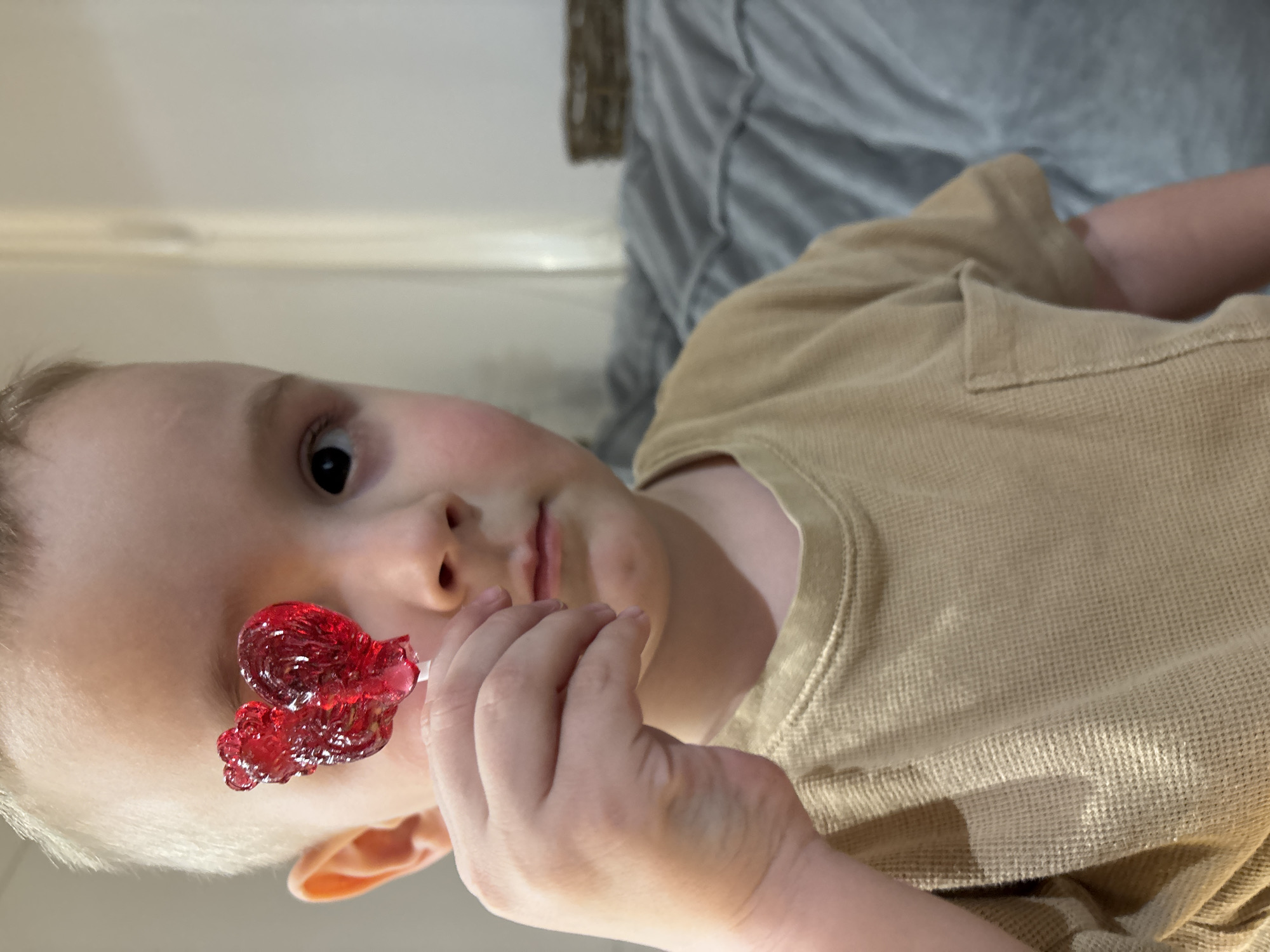 A young child with blonde hair is holding a red, heart-shaped lollipop. The child is wearing a tan-colored waffle-knit shirt. The background is out of focus, showing a light gray surface and a hint of white wall. The location is not discernible from the image.