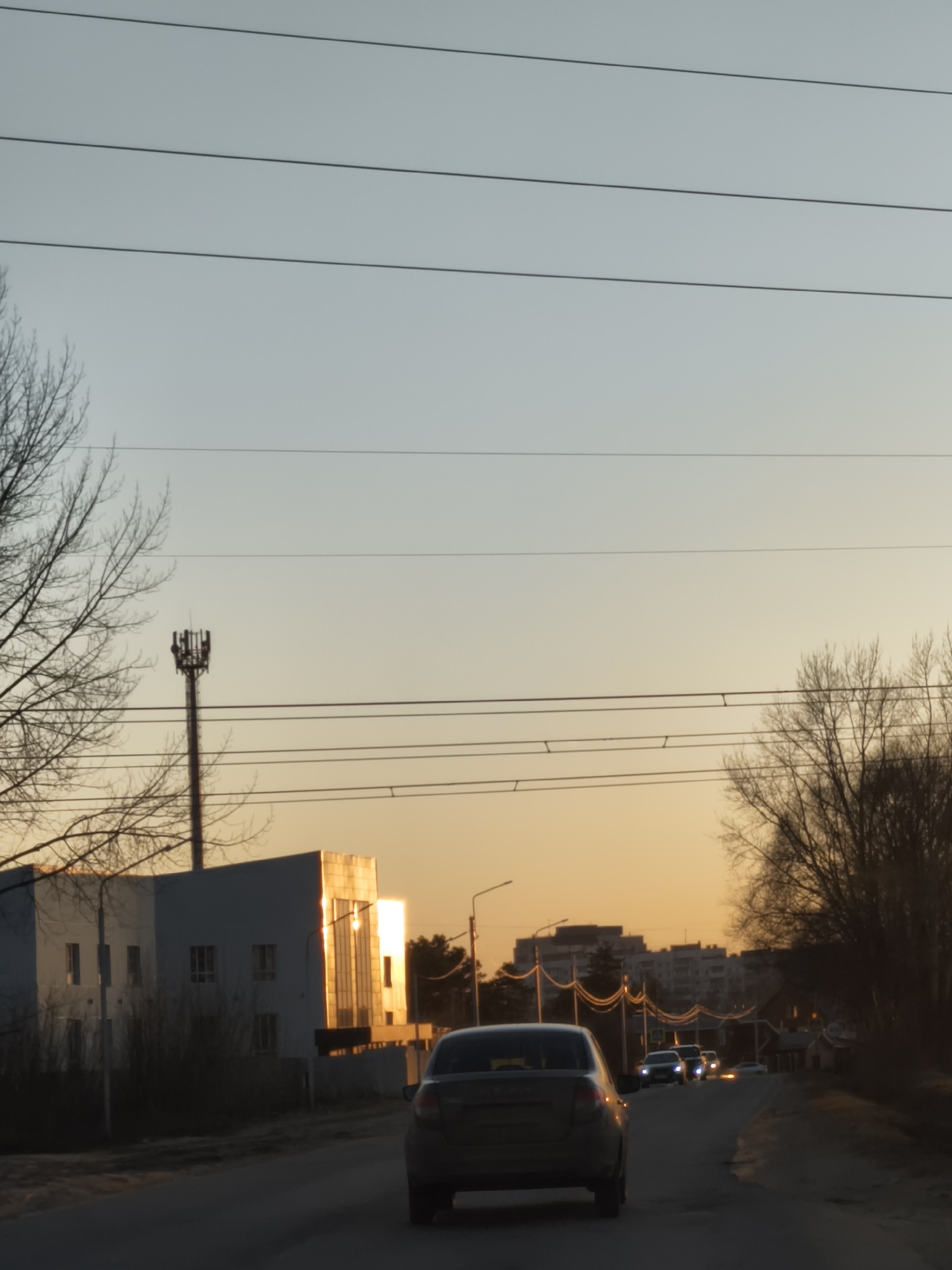 The image shows a road with several cars driving on it. A building is on the left side of the road, and trees are on both sides. A communications tower is next to the building on the left side. Overhead power lines are visible in the sky, which has a gradient from blue to orange. The scene appears to be at sunset. The location is Unknown, Unknown.