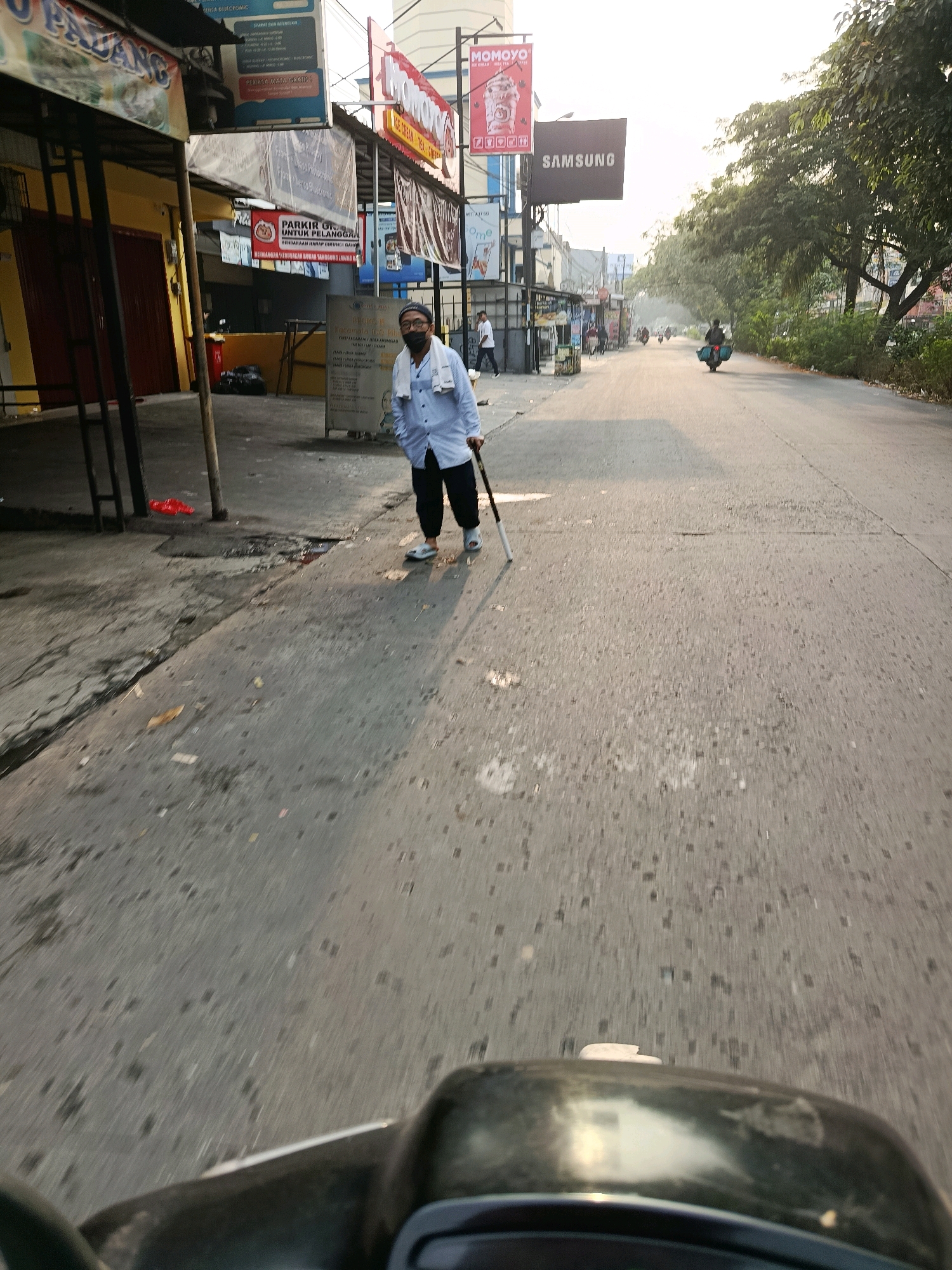 The image depicts a street scene in the city of Bekasi, Indonesia, likely during the morning or late afternoon given the low angle of the sun and long shadows. A man wearing a light blue long-sleeved shirt, dark pants, a face mask, and glasses is standing in the middle of the road, holding a white cane with a black tip. He is facing the camera and appears to be walking or about to walk. His stance suggests he might be visually impaired.
The road surface is asphalt, showing signs of wear and tear with cracks and scattered debris. To the left of the road, there are several storefronts with awnings and signage, some of which are in Indonesian. One sign visible from a closer crop reads "MOMOYO" with an image of an ice cream cone. Another sign displays the Samsung logo. A few motorcycles are visible on the road in the distance, with one in the middle of the frame carrying a passenger. Trees line the right side of the road, their leaves creating a canopy that casts shadows on the pavement. The overall atmosphere is that of a commercial street in a bustling Indonesian city. The foreground of the image shows a portion of a dark vehicle, possibly a scooter or motorcycle, from which the photo appears to have been taken.