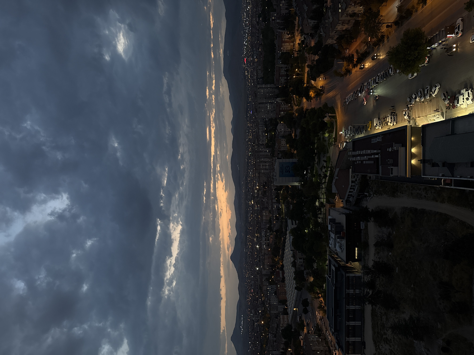 This is a high-angle, medium-shot photograph taken at dusk overlooking the city of Konya, Turkey. The sky is filled with dark, dramatic clouds with hints of orange and yellow light breaking through near the horizon, suggesting either sunrise or sunset. Below the sky, a panoramic view of the city unfolds, characterized by a dense collection of buildings illuminated by streetlights and interior lights. In the foreground, several buildings are visible, including a large, dark-colored structure with an illuminated window. To the right, a parking lot is filled with numerous cars, with a few people visible walking near them. A street with cars driving on it runs alongside the parking lot, with streetlights illuminating the scene. In the distance, mountains form a silhouetted backdrop to the city, with scattered lights indicating further settlements. The overall atmosphere is twilight urban.