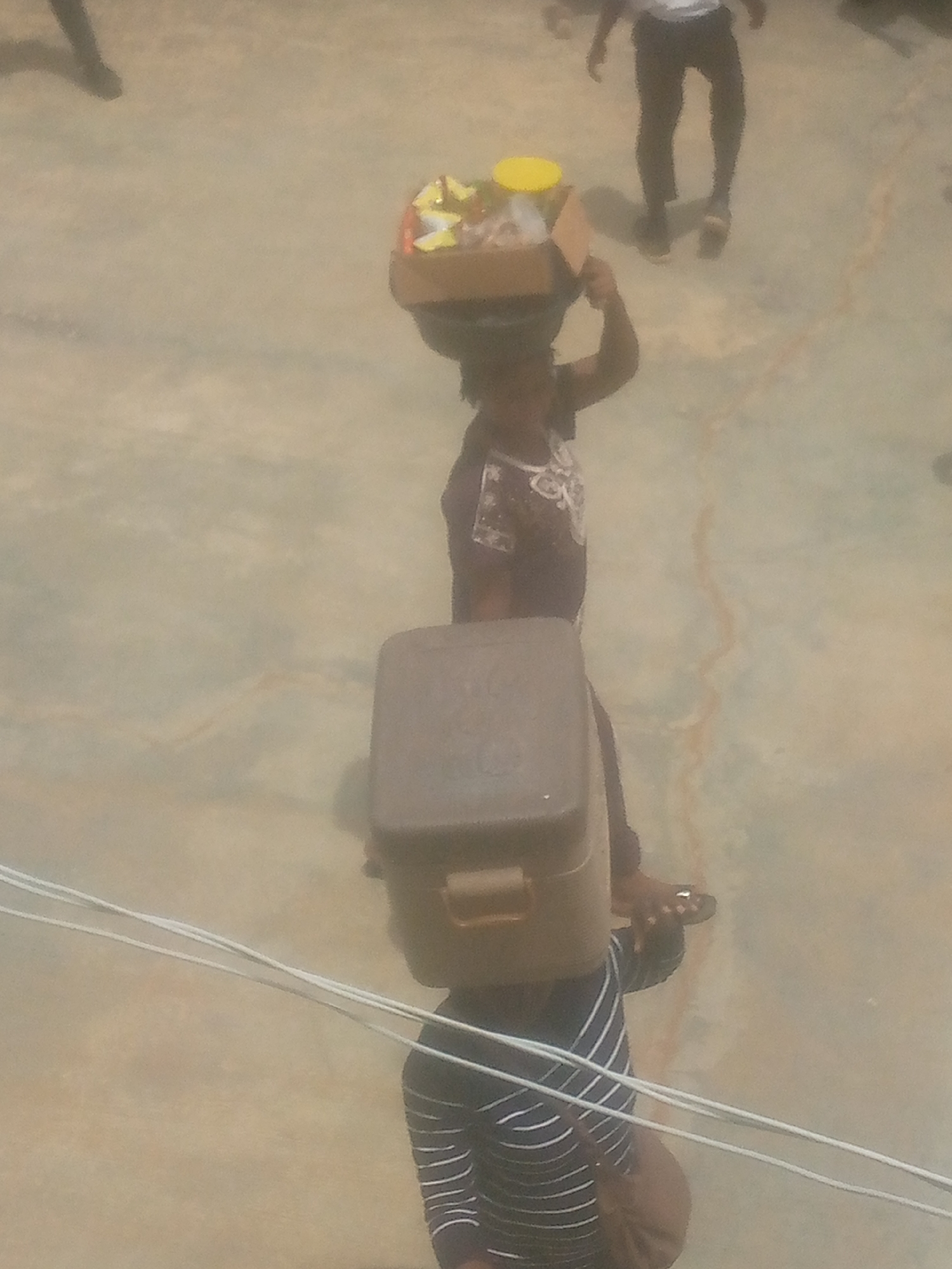 The image shows two individuals in Ikorodu, Nigeria, both carrying items on their heads.
The person closer to the foreground wears a striped shirt and carries a brown, rectangular cooler. The second individual is slightly behind, wearing a patterned shirt, and balancing a box containing food items on their head. A third person is walking out of frame towards the background. The background surface has a mottled appearance. Three white cables cut across the lower part of the image.