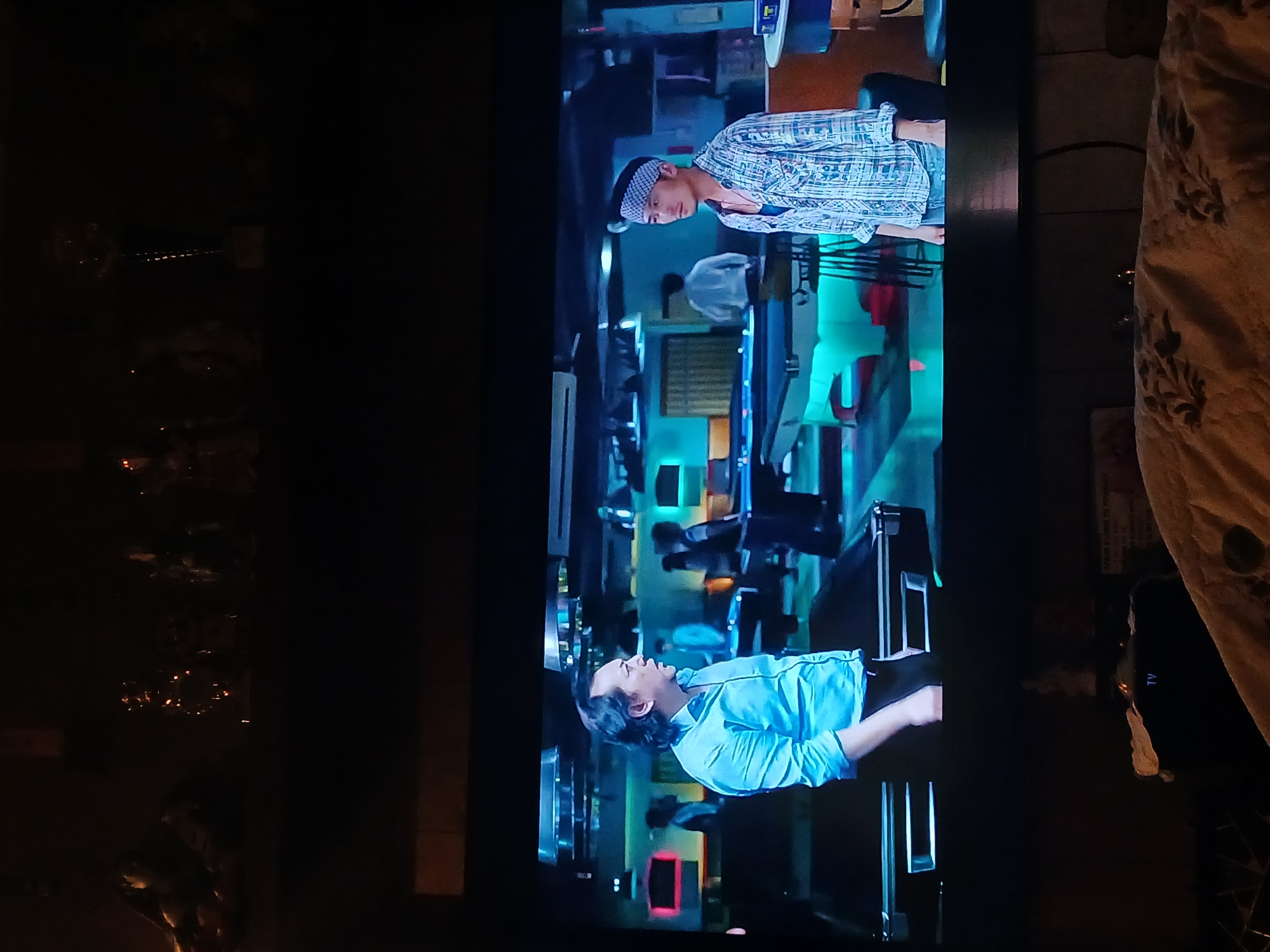 The image depicts a television screen displaying a scene with two men in what appears to be a restaurant setting. The man on the left side of the screen is wearing a light blue shirt and has dark hair. The man on the right side of the screen is wearing a patterned blue and white shirt and a matching headband. The restaurant interior features a mix of blue and green lighting with tables and chairs visible in the background. The photograph appears to have been taken in Bengaluru, India. The location is inferred from the context and is not explicit within the image itself.