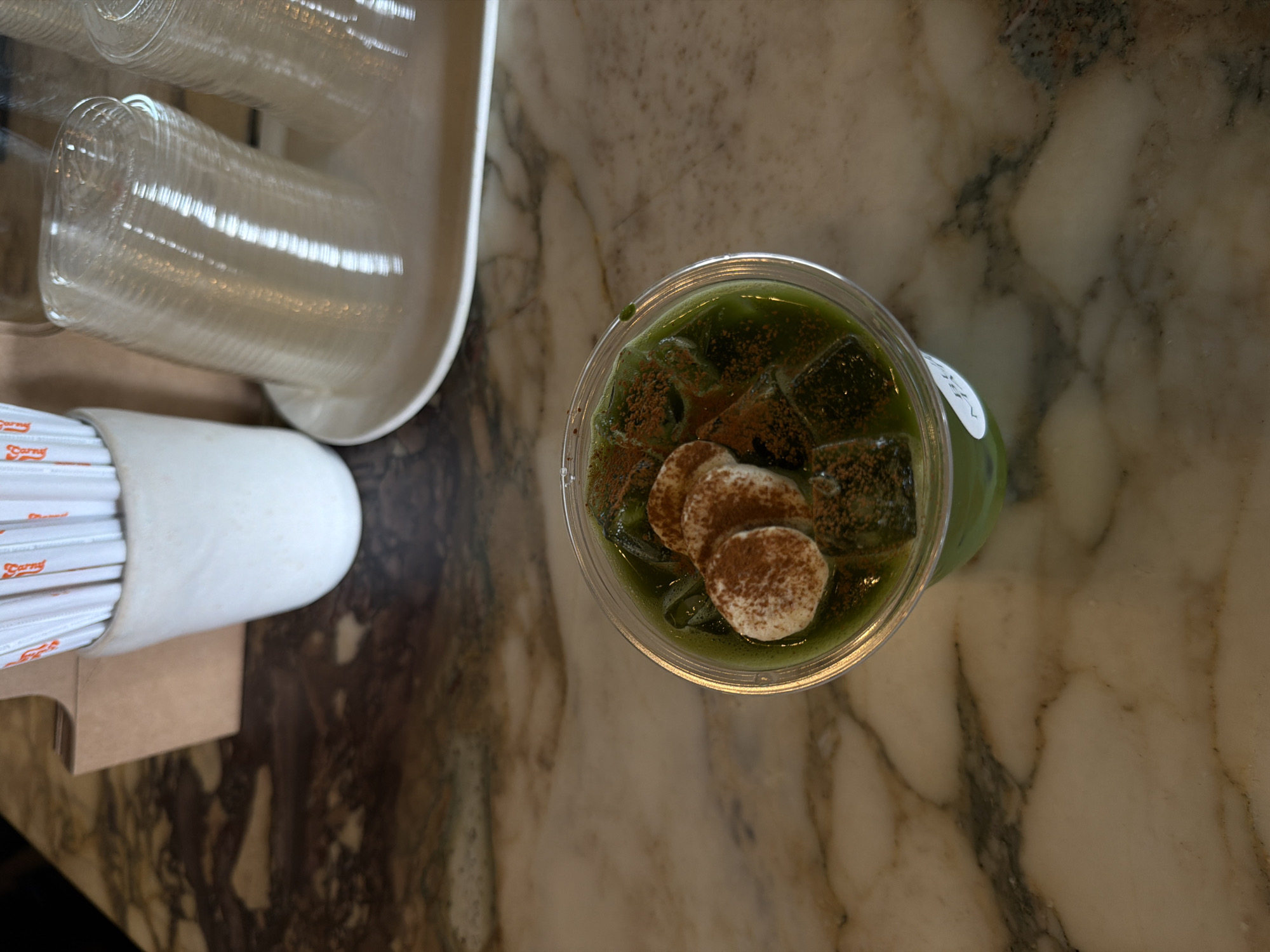 The image, taken from an overhead perspective, showcases a clear plastic cup containing a green beverage topped with ice cubes and three circular marshmallows dusted with a brown powder, likely cocoa. This drink sits on a marble countertop.
To the left of the cup are a dispenser containing clear plastic cups and a dispenser filled with white straws. The items are arranged on a countertop.
Based on the branding visible on the straw wrappers, the location is likely a Carns Palm Desert store. Palm Desert is a city located in the United States of America.