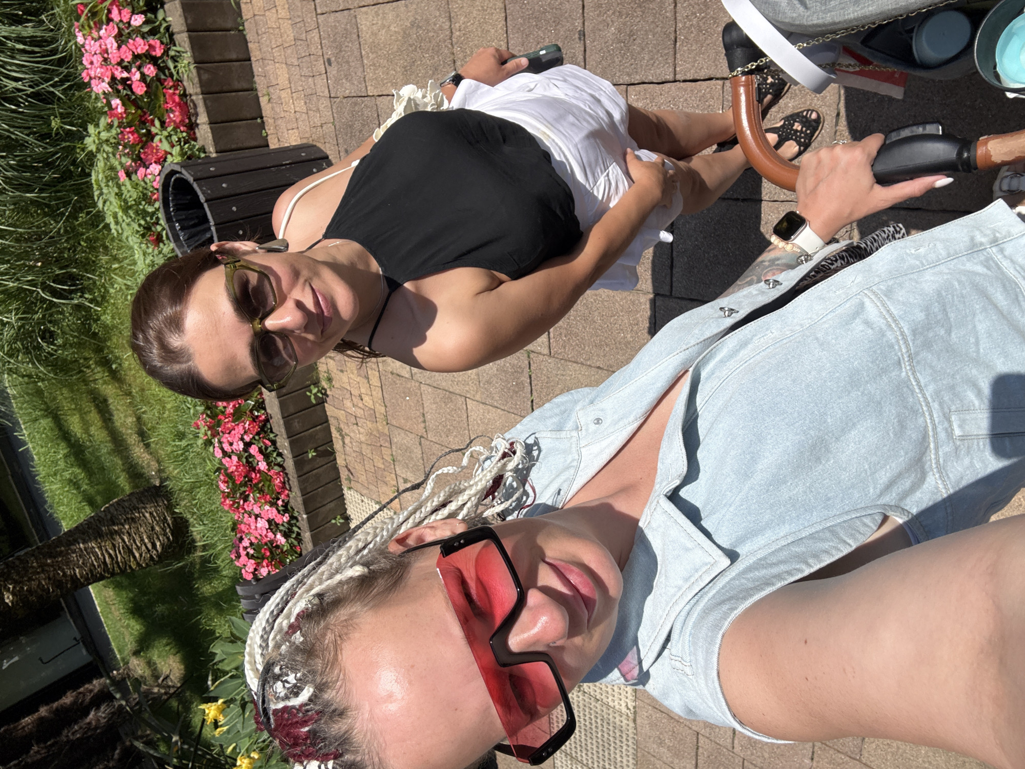 The image shows two women outdoors on a sunny day, likely in a park or public walkway in Sochi, Russia. The woman in the foreground, on the left, has blonde hair styled in braids with red and grey streaks and is wearing large, angular red-tinted sunglasses and a light blue denim vest. She is taking a selfie. The woman in the background, on the right, has brown hair, is wearing green-tinted sunglasses, a black halter top, and a white skirt. She is smiling at the camera. In the background, there is a flowerbed with pink and red flowers, green grass, and a palm tree. A black trash can is visible behind the woman in the background. The ground is paved with interlocking bricks. A stroller is partially visible in the lower right corner of the image.