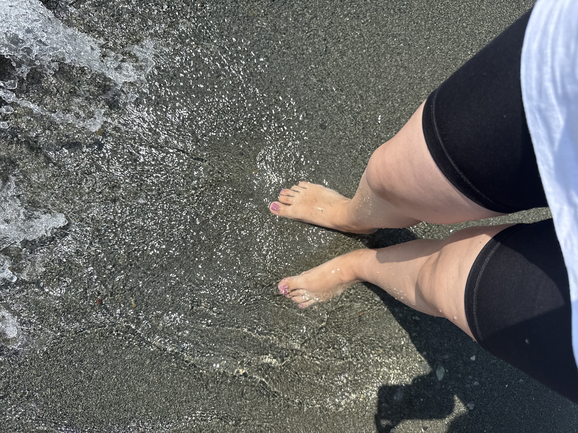 The image shows a top-down view of a person's legs and feet partially submerged in shallow, clear water on a dark, pebbly beach. The water is breaking into small waves at the top of the frame, creating a sparkling effect. The person is wearing black shorts and a white shirt is visible at the bottom. There is a shadow of a person or object on the left side of the frame. The scene appears to be a natural body of water, possibly a lake or the sea, in an outdoor environment. The location is specified as Tsamakaberd, Armenia.