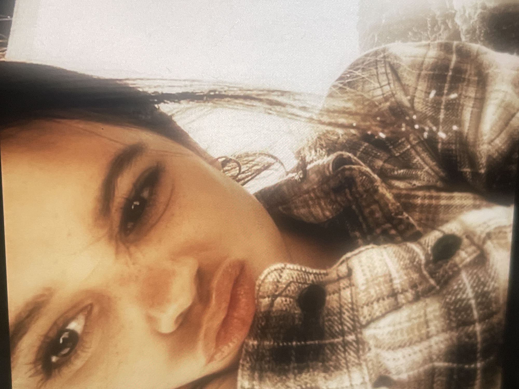 A close-up, angled portrait features a young woman with dark hair and striking dark eyes, looking directly at the viewer. Her face, characterized by freckles around her nose and cheeks, dominates the left side of the frame. She has full lips, slightly parted, with a subtle, perhaps pouty or intense, expression. Her dark hair frames her face, and her eyebrows are prominent.
She is wearing a plaid shirt, visible on the right side and across her shoulder. The shirt features a checkered pattern in earthy tones of brown and beige, and buttons are discernable on the fabric. The texture of the shirt suggests it might be made of flannel or a similar soft, warm material.
The background is largely out of focus and overexposed, appearing bright white towards the top, with some darker, blurred shapes hinting at an indistinct environment behind her. The overall lighting suggests a bright setting, though specific time of day or weather cannot be definitively determined due to the blurred and bright background. The image has a slightly warm, sepia-toned cast and shows signs of digital artifacts or being a photograph of a screen, contributing to a slightly grainy appearance. There is no visible text. The image does not contain any visual cues to confirm the location in Poltava, Ukraine.