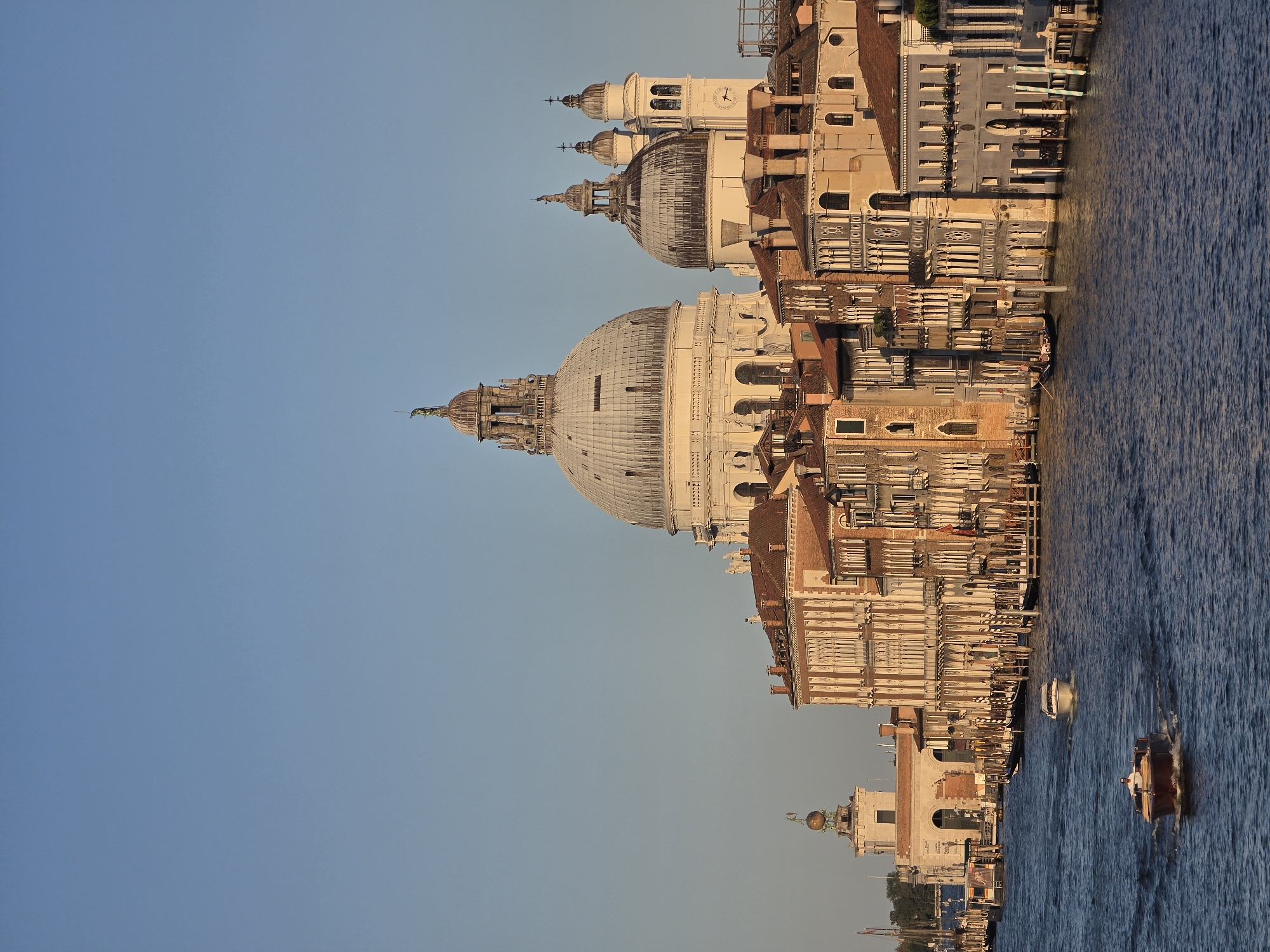 The image depicts a view of the Grand Canal in Venice, Italy, with the Basilica di Santa Maria della Salute prominently featured in the background. The architecture of Venice, characterized by its historic palazzi and canals, is evident. Several boats are visible on the water, including what appears to be a water taxi in the foreground with a person at the helm. Another smaller white boat is further in the distance. Gondolas are also present along the canal. The sky is clear and blue, suggesting a daytime scene, likely during the late afternoon or early evening given the warm lighting on the buildings. The water of the canal is a deep blue, reflecting the surrounding structures and sky. The buildings lining the canal exhibit ornate facades, typical of Venetian Renaissance and Baroque styles, with tiled roofs and numerous windows. The overall atmosphere is one of serene urban maritime life.