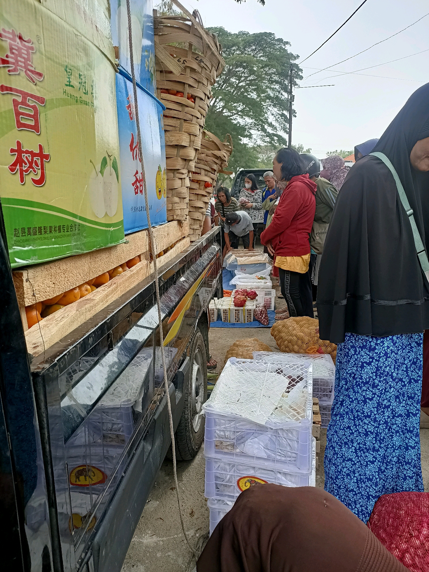 A street market in Madiun, Indonesia, features a truck loaded with fruit boxes and baskets. In the foreground, a person wearing a blue floral skirt and a black hijab is partially visible. To the left, a black truck bed is laden with a green box displaying Chinese characters and images of pears, and a blue box with Chinese text and cartoonish fruit faces. Beneath these boxes, several rows of oranges are visible. A large wicker basket filled with fruit is stacked on top of the boxes. Behind the truck, several people are gathered, some appearing to be shoppers or vendors. The ground is paved with concrete. The scene suggests a bustling commerce environment, possibly during the morning or afternoon given the ambient light.