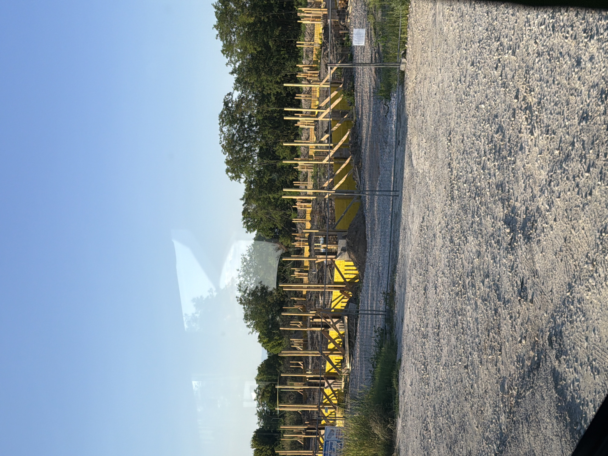 The image shows a construction site in Tsaratsovo, Bulgaria. The foreground is a gravel surface with tire tracks. In the midground, a construction site is visible with yellow formwork and wooden supports for what appears to be a foundation. Numerous vertical wooden posts are erected throughout the site. A chain-link fence runs across the midground, separating the viewer from the construction area. In the background, a line of green trees forms a backdrop under a clear blue sky. There are no people visible in the image.