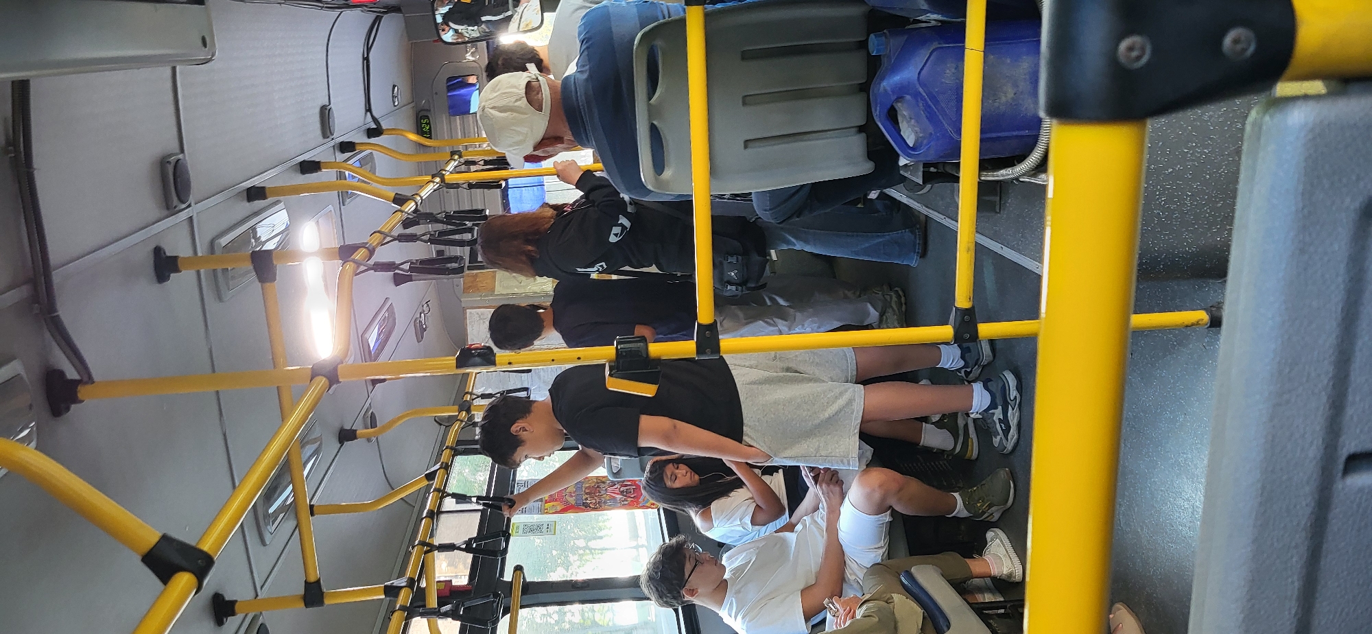 Inside a bus, several individuals are visible, some seated and others standing. The bus interior features prominent yellow handrails and poles, with grey seats. A blue plastic container is placed on the floor in the foreground. The scene appears to be on a bus in Oskemen, Kazakhstan, as indicated by the bus's interior configuration and passenger demographics, which are consistent with public transportation in that region.