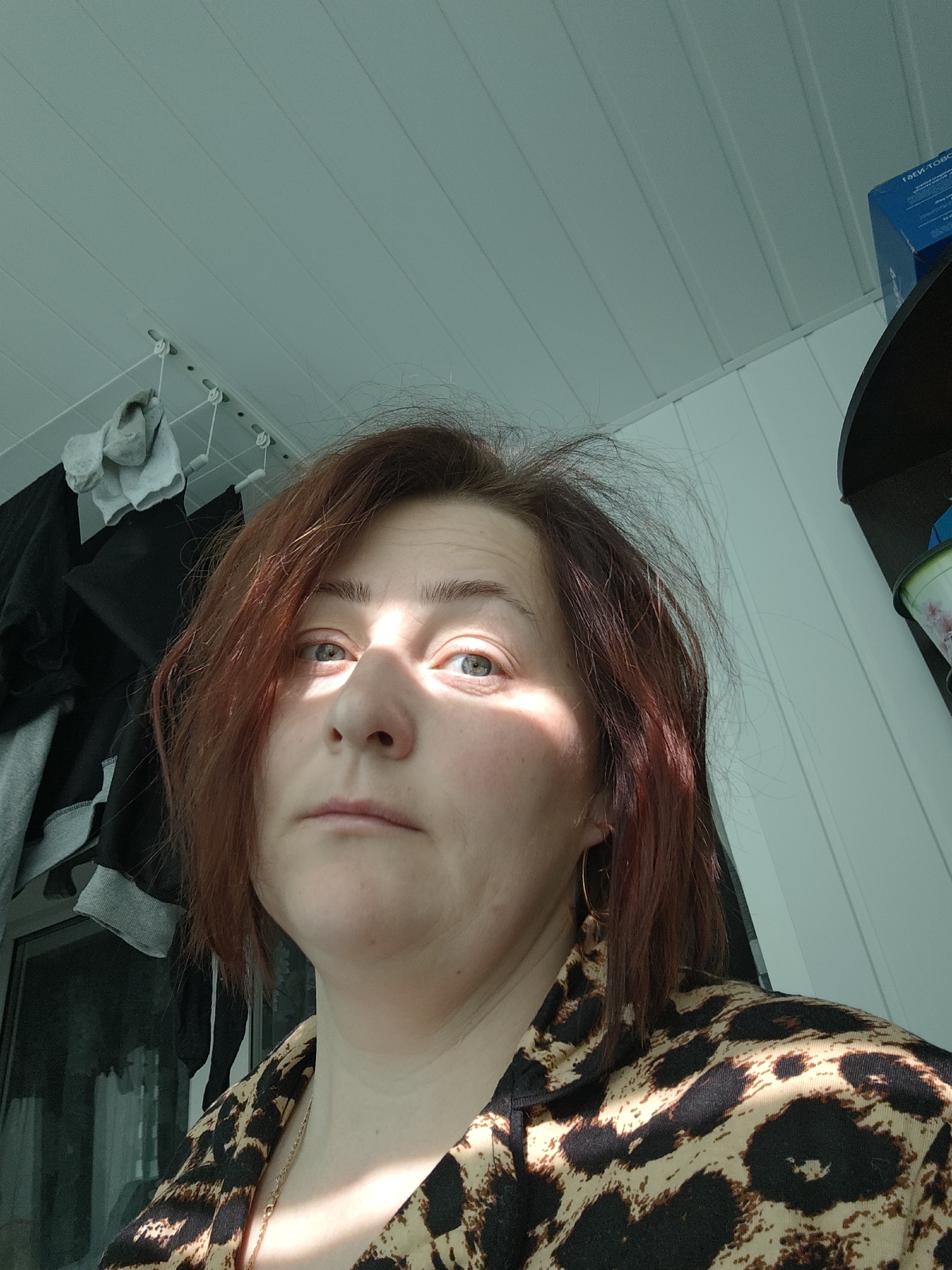 A woman with red-brown hair, wearing a leopard-print shirt and gold hoop earrings, is shown in a medium close-up shot from a low angle. Her face is partially illuminated by sunlight, creating strong shadows. A clothes drying rack with clothes hanging on it is visible in the background to her left. A dark shelf with a blue box on it is visible to her right. The ceiling above is paneled. The location cannot be determined from the image.