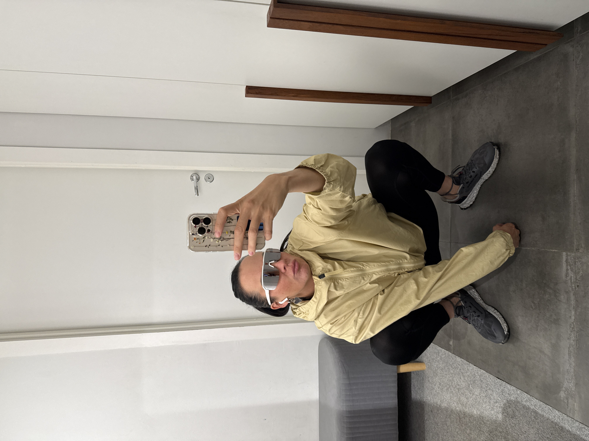 A woman with dark hair pulled back into a ponytail is crouching in a room, taking a selfie with her phone. She is wearing white futuristic sunglasses, a light yellow windbreaker jacket zipped up, black leggings, and dark grey athletic shoes with white speckles on the soles. She has on small hoop earrings with a heart-shaped charm. In the background, to her left, is a grey upholstered bench with wooden legs. To her right, there are white doors with a silver door handle and a white frame, and further to the right, two vertical wooden posts are visible. The floor is tiled in large grey squares. The overall environment appears to be an indoor dressing room or a similar functional space within a residential or commercial building, potentially located in Chaikovsky, Russia.