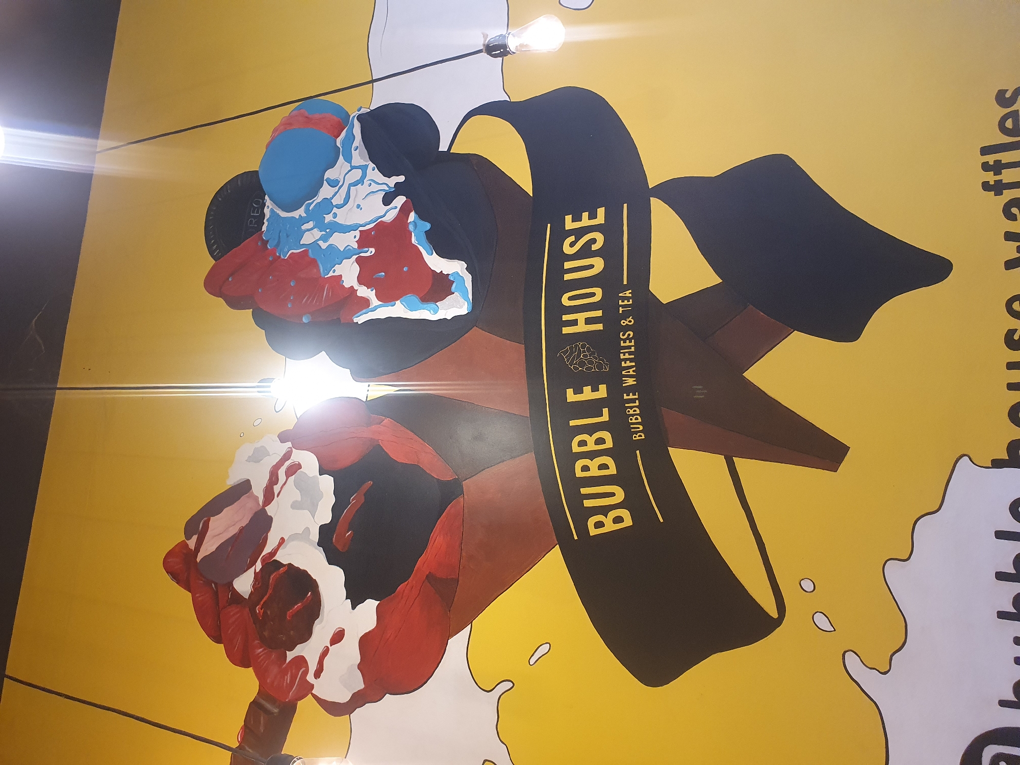 This media file captures a vibrant, brightly lit indoor mural, likely part of a dessert or cafe shop in Plovdiv, Bulgaria. The mural, set against a cheerful yellow background, features two large, stylized bubble waffle cones, each held by a cartoonish brown arm.
The waffle cone on the right is topped with a colorful mound of blue and red, possibly ice cream or whipped cream, adorned with white and light blue drizzles. A dark, round object, partially revealing the brand name "OREO," is nestled among the toppings. The waffle cone on the left is generously topped with white, likely whipped cream, and rich red drizzles, suggesting a berry or cherry sauce.
Abstract white splatters and shapes are scattered across the yellow wall, mimicking dessert drips and adding a playful element to the design. Two bare incandescent light bulbs, hanging from thin wires, are visible, casting bright lens flares on the image and indicating artificial indoor lighting.
Prominently displayed on a black banner running vertically through the center of the mural is the text "BUBBLE HOUSE" in large yellow capital letters. Below this, in smaller yellow text, it specifies the offerings: "BUBBLE WAFFLES & TEA". On the far right edge, partially cropped, further text consistent with a social media handle or website name is visible, reinforcing the brand. The overall scene creates an inviting and whimsical atmosphere for a dessert establishment.