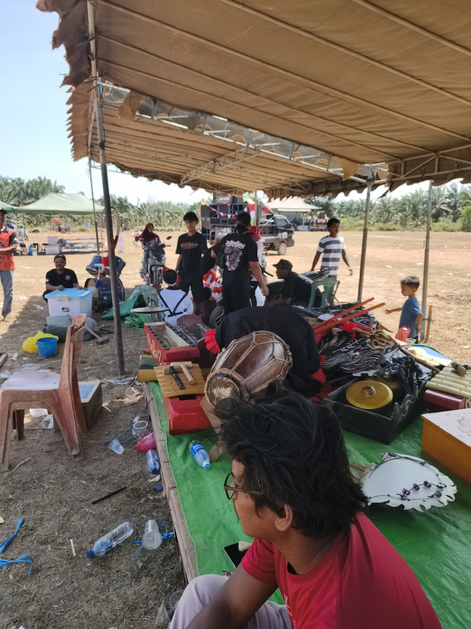 The image depicts an outdoor scene at an event in Pelanjau Jaya, Indonesia, under a large, tan canopy. In the foreground, a person wearing a red shirt is seated, facing away from the camera. In front of them is a table covered with a green tarp, displaying various musical instruments, including what appears to be a gamelan set with wooden bars and a drum. Several water bottles are scattered on the ground around the table.
Further back, under the canopy, several individuals are visible. Some are standing and interacting, while others are seated. Musical equipment, including speakers and what looks like a mixing board, is set up. In the background, there are glimpses of palm trees, indicative of a rural or semi-rural setting. A motorcycle is parked near some tents in the middle ground, and a few people are gathered around it. The ground is covered in dry grass and dirt. The overall atmosphere suggests a preparation or break period during an outdoor performance or gathering.
