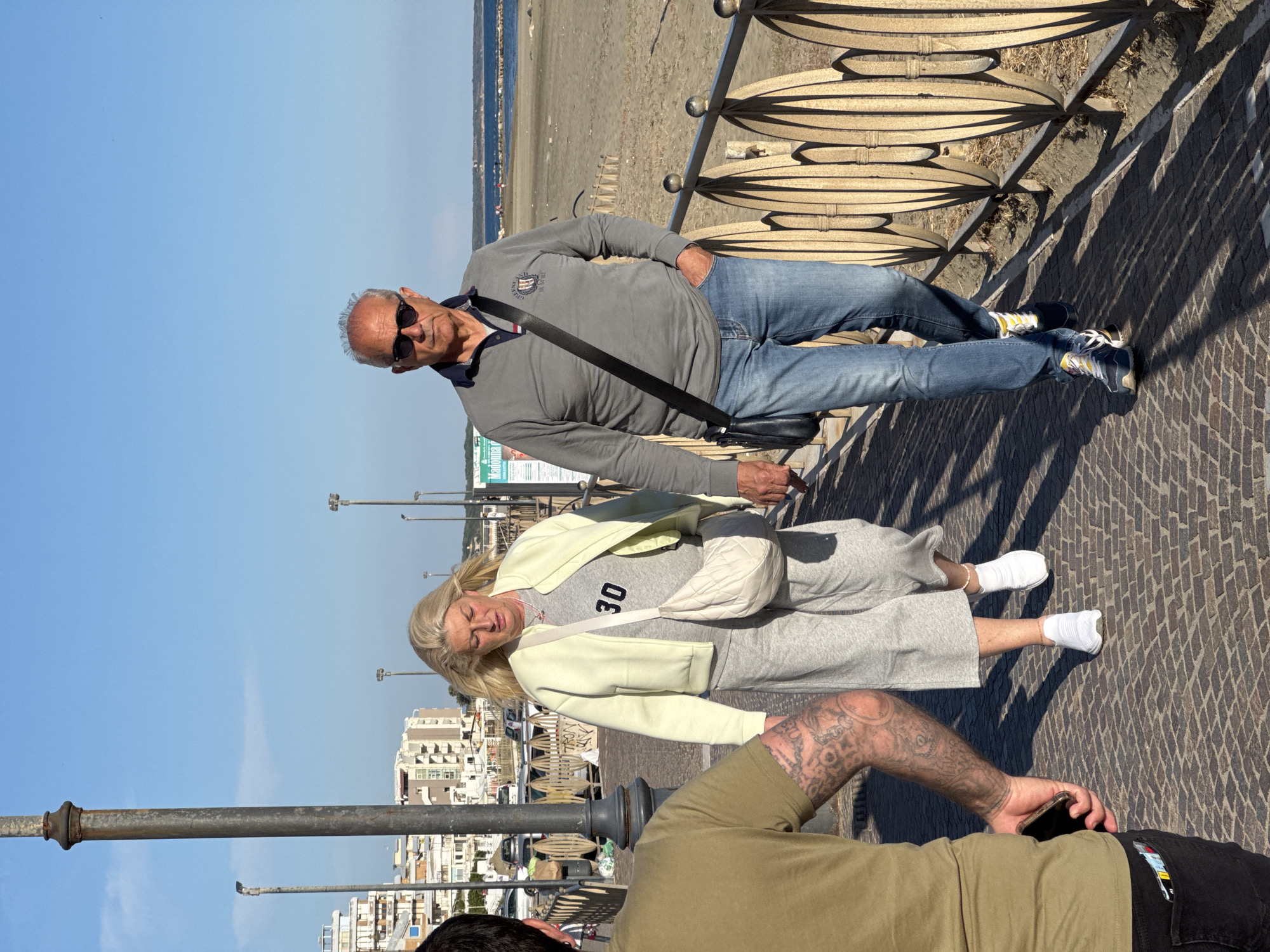 This image captures a bright, sunny scene on a promenade in Nettuno, Italy. The photo is rotated, showing the horizon vertically. In the middle ground, an older man and woman are featured. The man, with grey hair and sunglasses, wears a grey long-sleeved polo shirt with an emblem, blue jeans, and athletic shoes with yellow accents. He stands with one hand in his pocket and a black shoulder bag across his body. The woman, with blonde hair, walks beside him, dressed in a light yellow jacket over a grey t-shirt with the number "30" printed on it, grey sweatpants, and white sneakers. She carries a light-colored quilted cross-body bag. Her expression is neutral, possibly squinting in the sun.
In the foreground (bottom of the rotated image), a person's tattooed arms, wearing an olive green shirt, are visible, holding a smartphone. Another person's legs in black pants are partially seen. The background reveals a wide, clear blue sky overhead, with a sandy beach and the sea beyond the promenade's decorative metal railing. In the distance, coastal buildings line the shore. Strong shadows on the patterned brick pavement indicate midday or early afternoon sunlight. A sign with partially visible text, possibly "Madonnar", is seen behind the man.