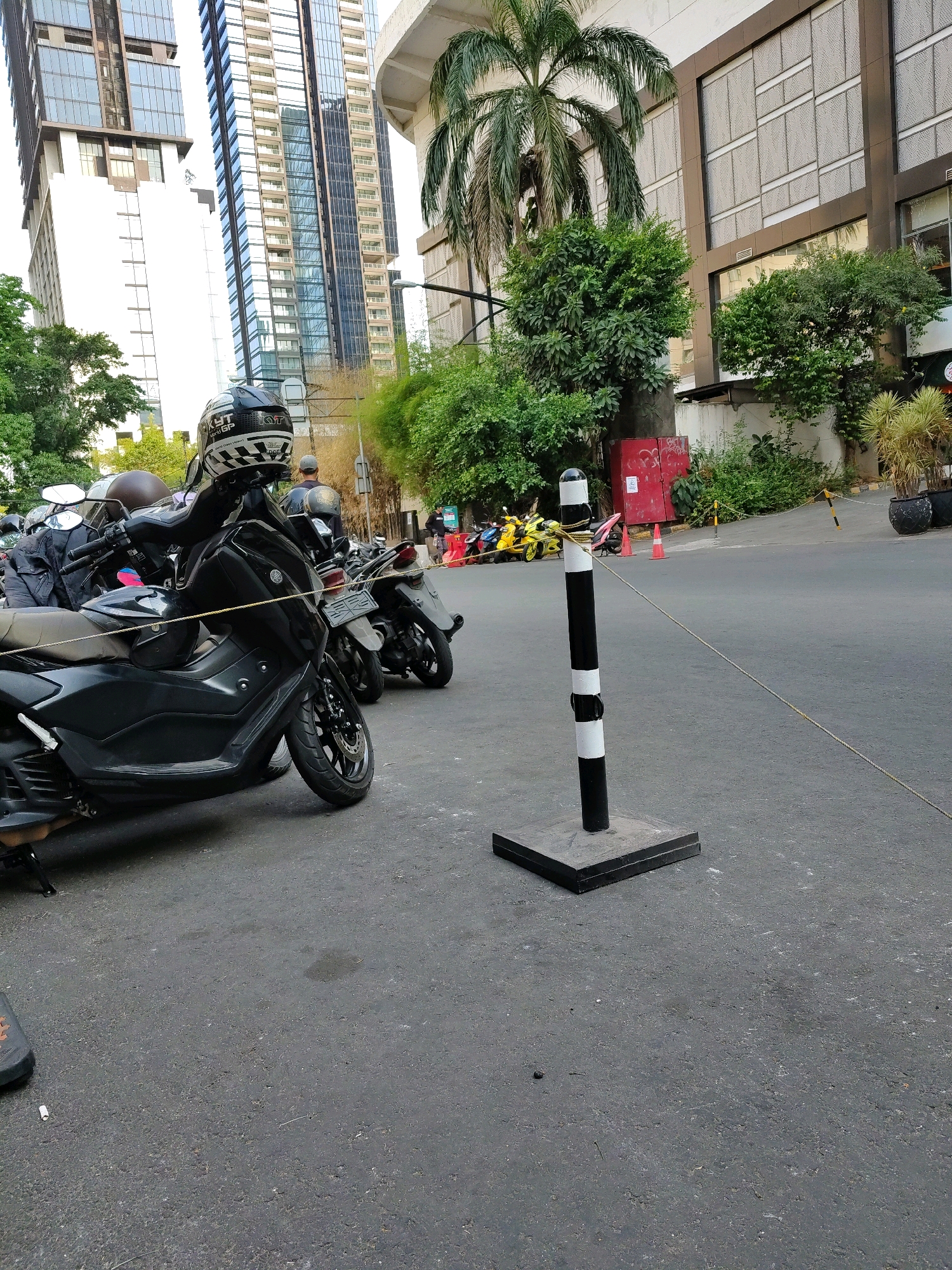 The image depicts an outdoor scene on a paved street in South Jakarta, Indonesia. In the foreground, a black scooter is parked, with its front wheel turned to the left. A black and white striped bollard with a black base stands to the right of the scooter, with a rope extending from it diagonally across the frame. Behind the scooter, other parked motorcycles are visible, including a yellow one further down the street. Tall, modern buildings with reflective windows dominate the background, interspersed with green trees and foliage. The sky is overcast. A red structure, possibly a utility box or small building, is visible amidst the greenery on the right side of the frame. The overall impression is of an urban environment with a focus on parked vehicles and contemporary architecture.