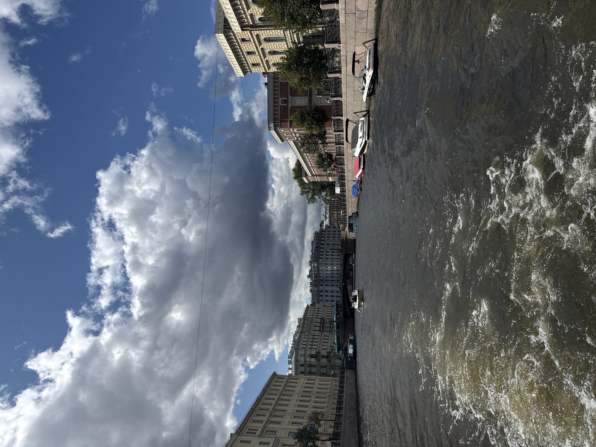 The image depicts a view from a moving boat on a canal in Saint Petersburg, Russia. The canal is lined with historical buildings and a stone embankment. Several boats are visible, including a white motorboat with two people in it in the middle of the canal, and several smaller boats docked along the embankment. The sky is partly cloudy with dramatic dark clouds interspersed with patches of blue sky. The water in the canal is dark and choppy, with a visible wake trailing behind the viewer's boat. The architecture of the buildings suggests a classical or neoclassical style common in Saint Petersburg. The overall scene conveys a sense of navigating through the city's waterways on a dynamic day.