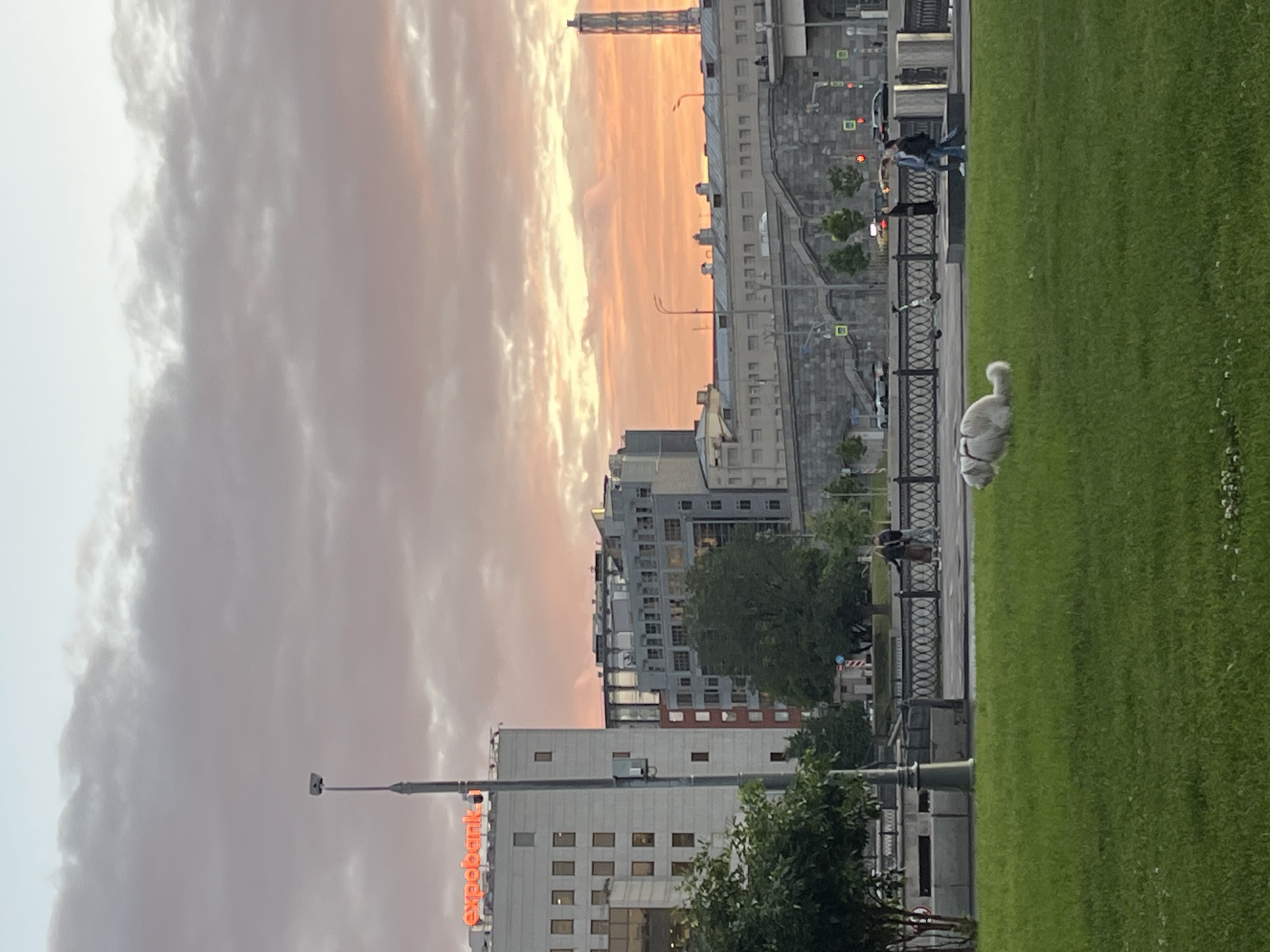 This is a vertical, slightly blurry image taken outdoors in what appears to be a city park. The scene depicts a fluffy white dog lying on a lush green lawn in the foreground. Behind the dog, a decorative black metal fence separates the park area from a sidewalk or pathway. On the pathway, two people are walking, one appearing to be holding onto a leash, suggesting they are with the dog, though the dog is not actively walking.
Beyond the pathway, a modern urban landscape is visible, characterized by tall buildings. The buildings have a mix of architectural styles, with some featuring many windows and balconies, and others appearing more monolithic. There are some trees scattered throughout the urban area, adding touches of green amidst the concrete structures. Traffic lights with red lights are visible, indicating ongoing traffic.
The sky above is filled with dramatic clouds, painted with soft hues of pink, orange, and white, suggesting it is either sunrise or sunset. This lends a peaceful and somewhat ethereal quality to the scene. The presence of streetlights and the general lighting suggest it is either early morning or late evening. A tall, slender structure, possibly a tower or a high-rise building, is visible in the distance against the colorful sky.
On one of the buildings in the mid-ground, the word "expobank" is visible in bright orange neon letters. The overall atmosphere is serene, with the focus on the peaceful coexistence of nature (the park) and urban life, with the dog and its owners enjoying the open space. The soft lighting and cloudy sky contribute to a calm and tranquil mood.