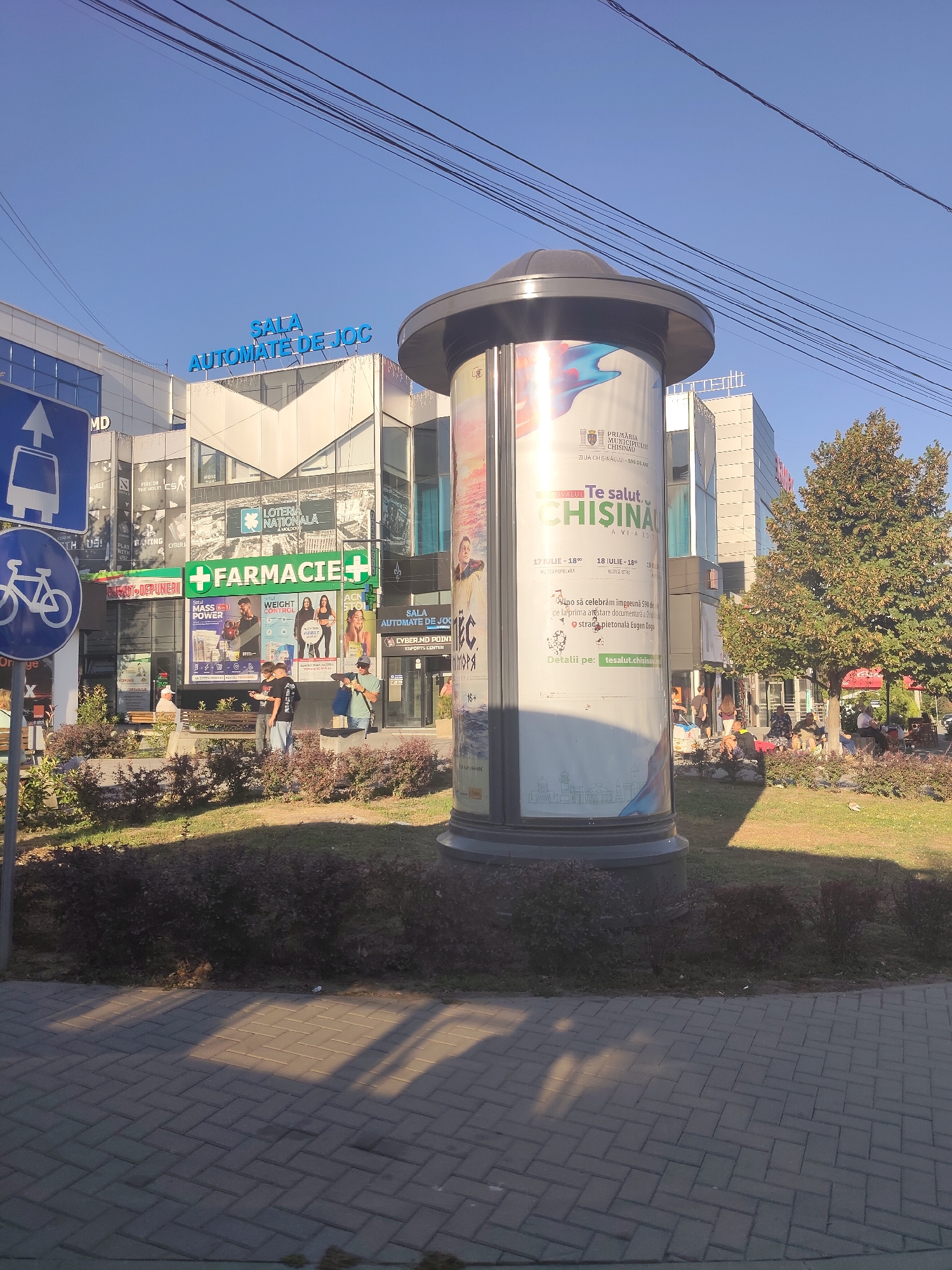 The image captures a street scene in Chișinău, Moldova, dominated by a modern building complex. A large, cylindrical billboard stands prominently in the foreground, displaying a colorful advertisement. To the left, a blue traffic sign with a white bus icon and a white bicycle icon indicates public transportation and cycling routes. Behind the billboard, the building features a sign reading "SALA AUTOMATE DE JOC" in bright blue letters. Various storefronts are visible, including one with a large green "FARMACIE+" sign. Several people are present, some walking and others seated on benches in a landscaped area in front of the building. The architecture suggests a commercial or entertainment venue, and the presence of the pharmacy and gaming hall points towards a mixed-use development. The overall environment is urban, with a clear blue sky and shadows cast by the sun suggesting a daytime setting.