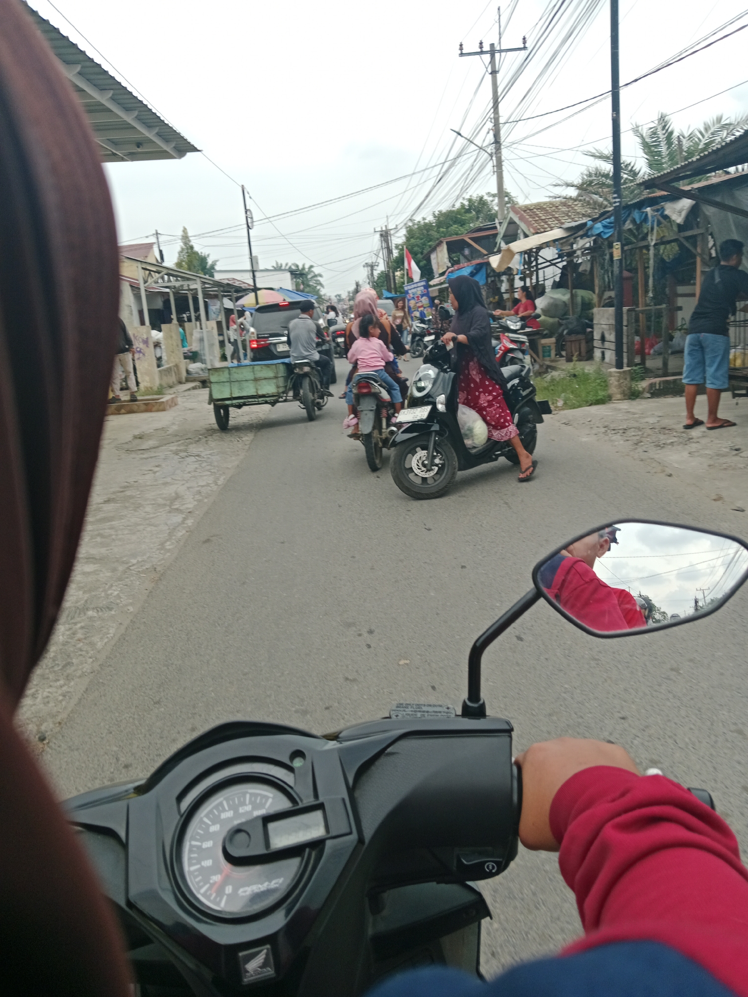 The image depicts a street scene in the City of Binjai, Indonesia, likely a busy marketplace area. A person wearing a brown hijab is riding a motorcycle, with their hand on the handlebar and their arm in a red sweatshirt visible in the foreground. A motorcycle mirror reflects the rider and the sky. In the midground, several other motorcycles are present, some carrying passengers. One motorcycle is pulling a small green trailer. People are seen walking and riding, with some standing near stalls on the side of the road. The environment suggests a commercial area with shops and makeshift structures lining the street. The sky is overcast.